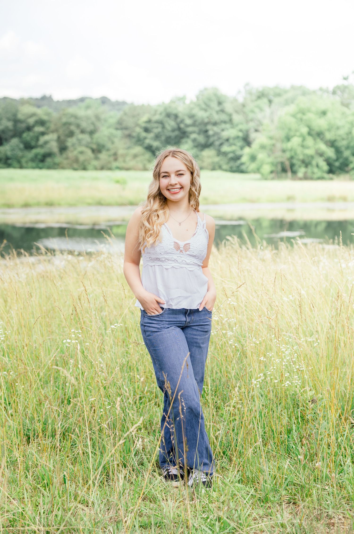 Young woman in casual attire poses in a golden field with a lake and trees in the background.