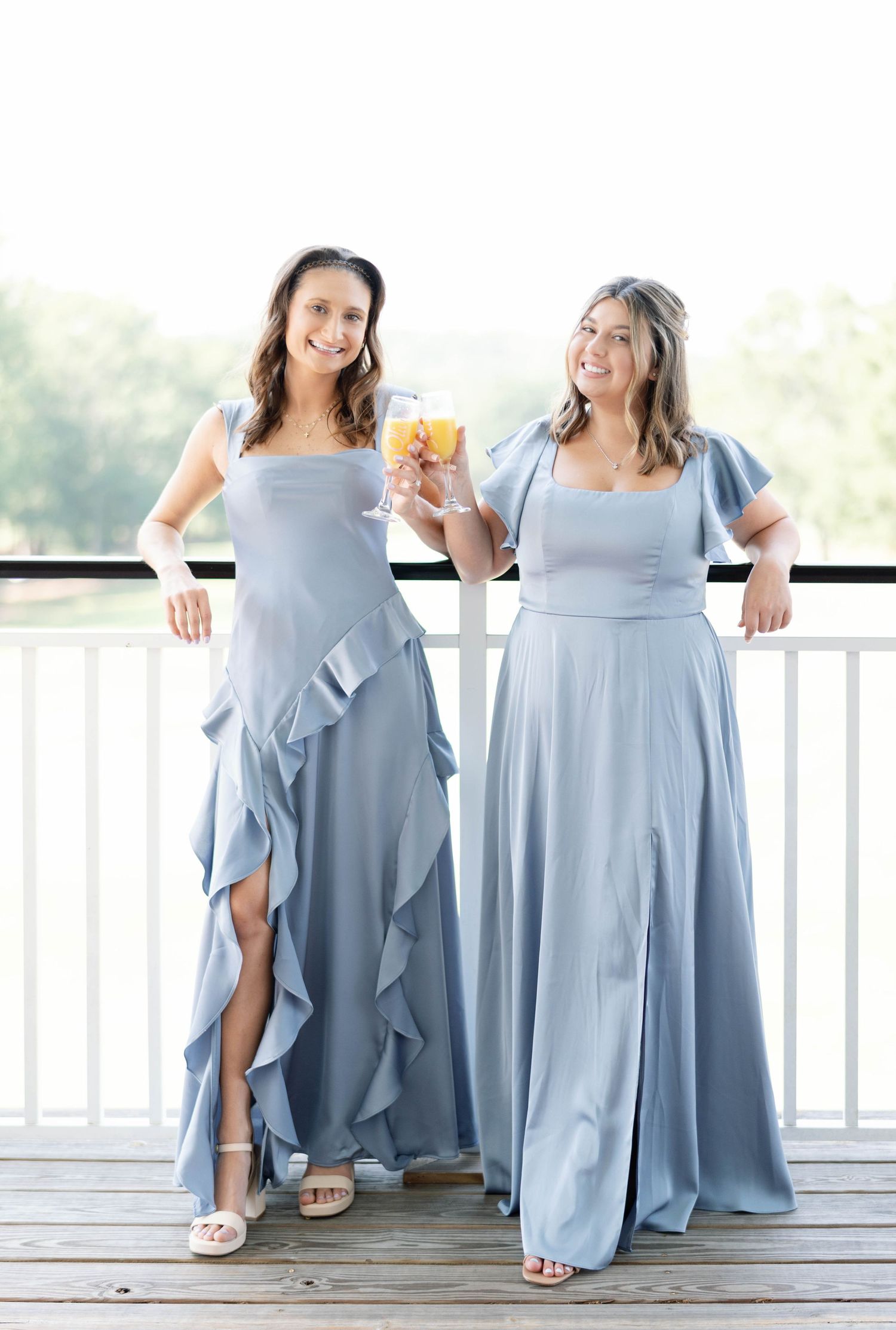 Bridesmaids in flowing dusty blue dresses pose together on a white porch railing during a wedding celebration.