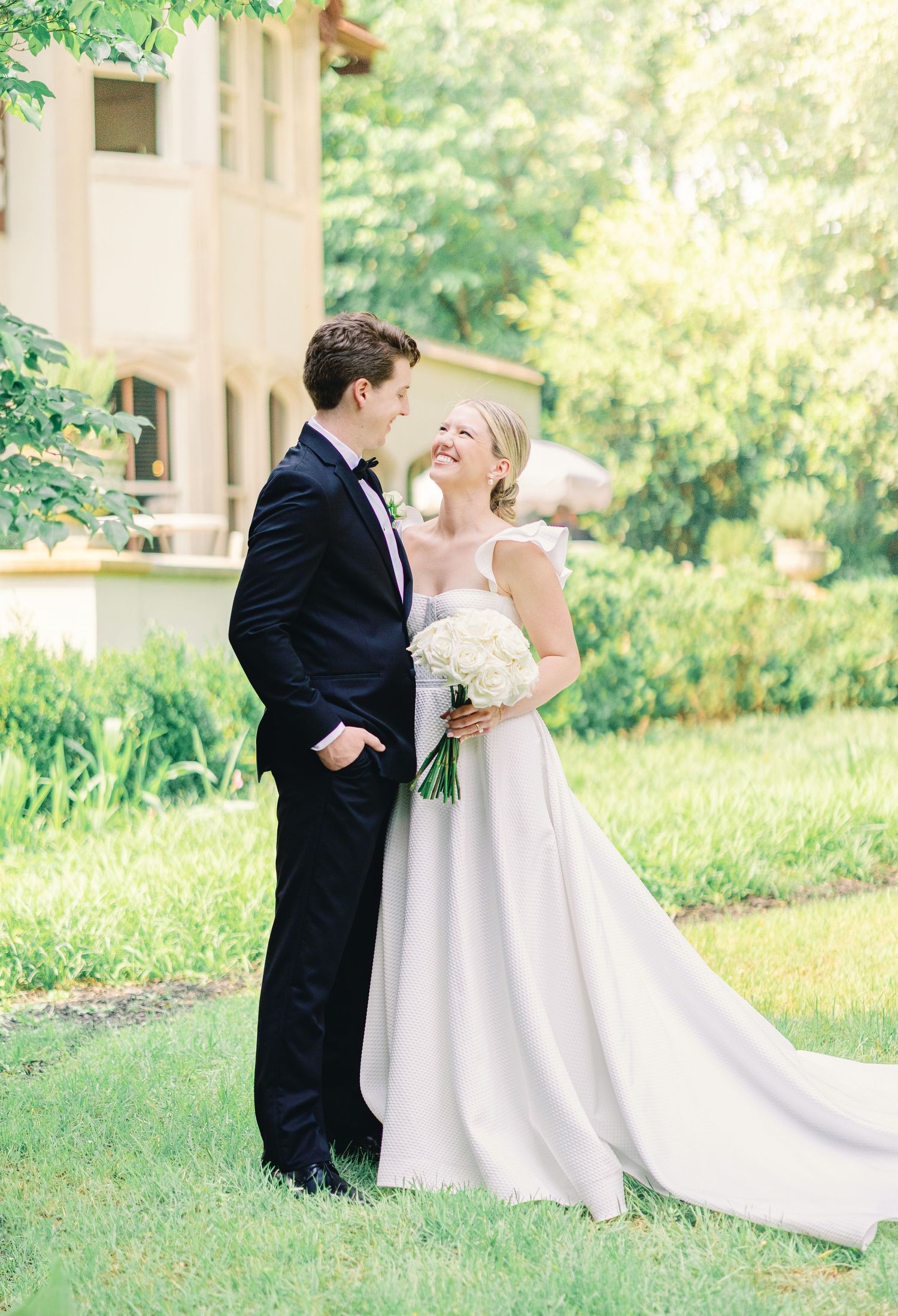 A couple shares a joyful moment in their wedding attire on a sunlit garden lawn beside a classic building exterior.