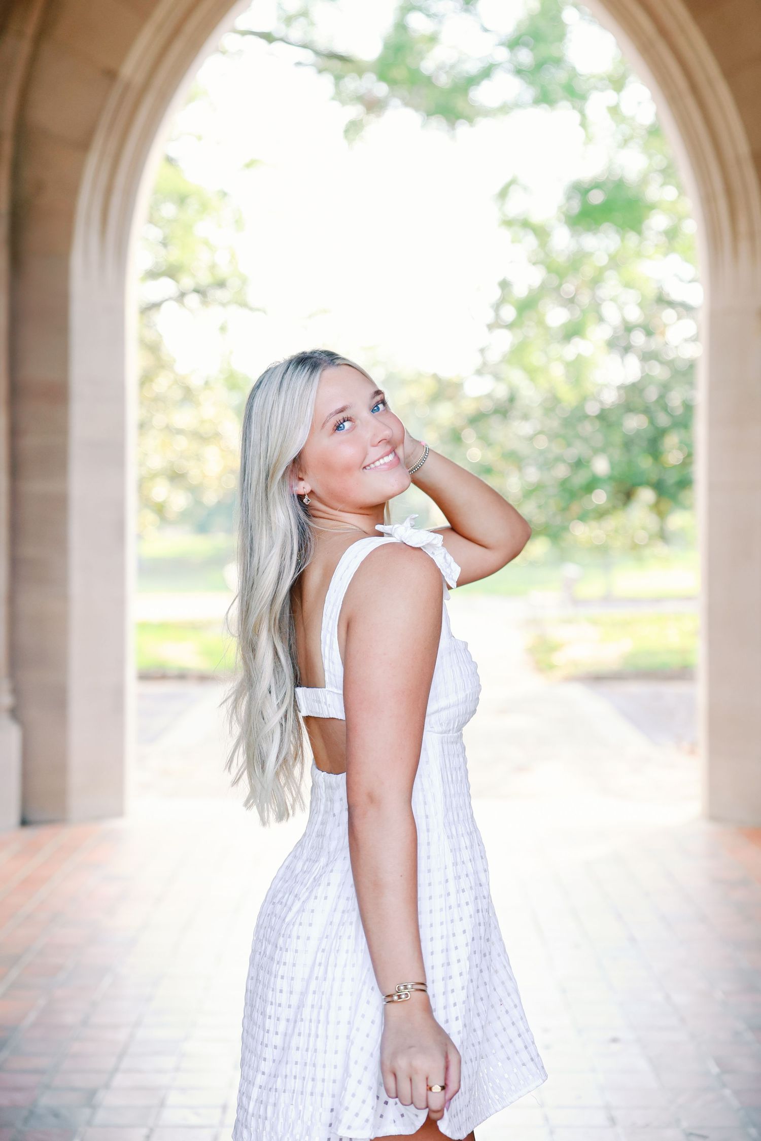 A person in a white eyelet summer dress poses under an arched walkway with sunlit foliage in the background.