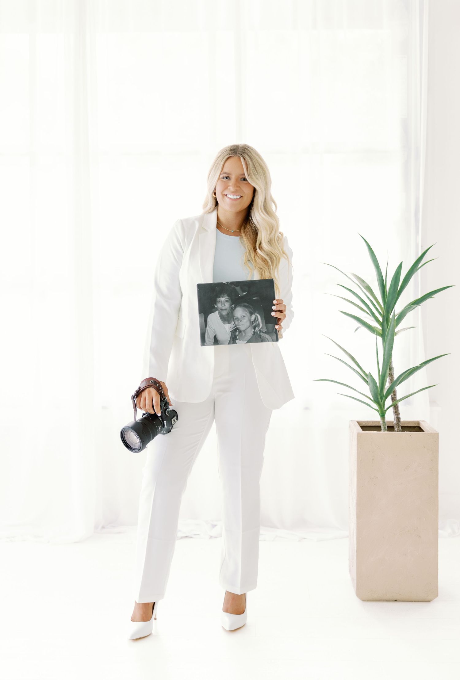 Fashionable figure in white outfit poses against bright background holding photo prints next to minimalist plant decor.