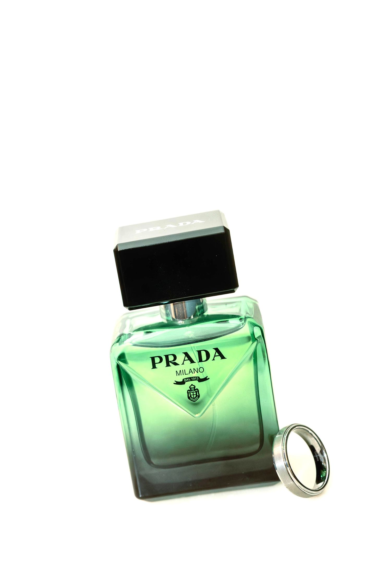 A green glass bottle of Prada perfume with a black cap sits beside a silver ring on a white background.