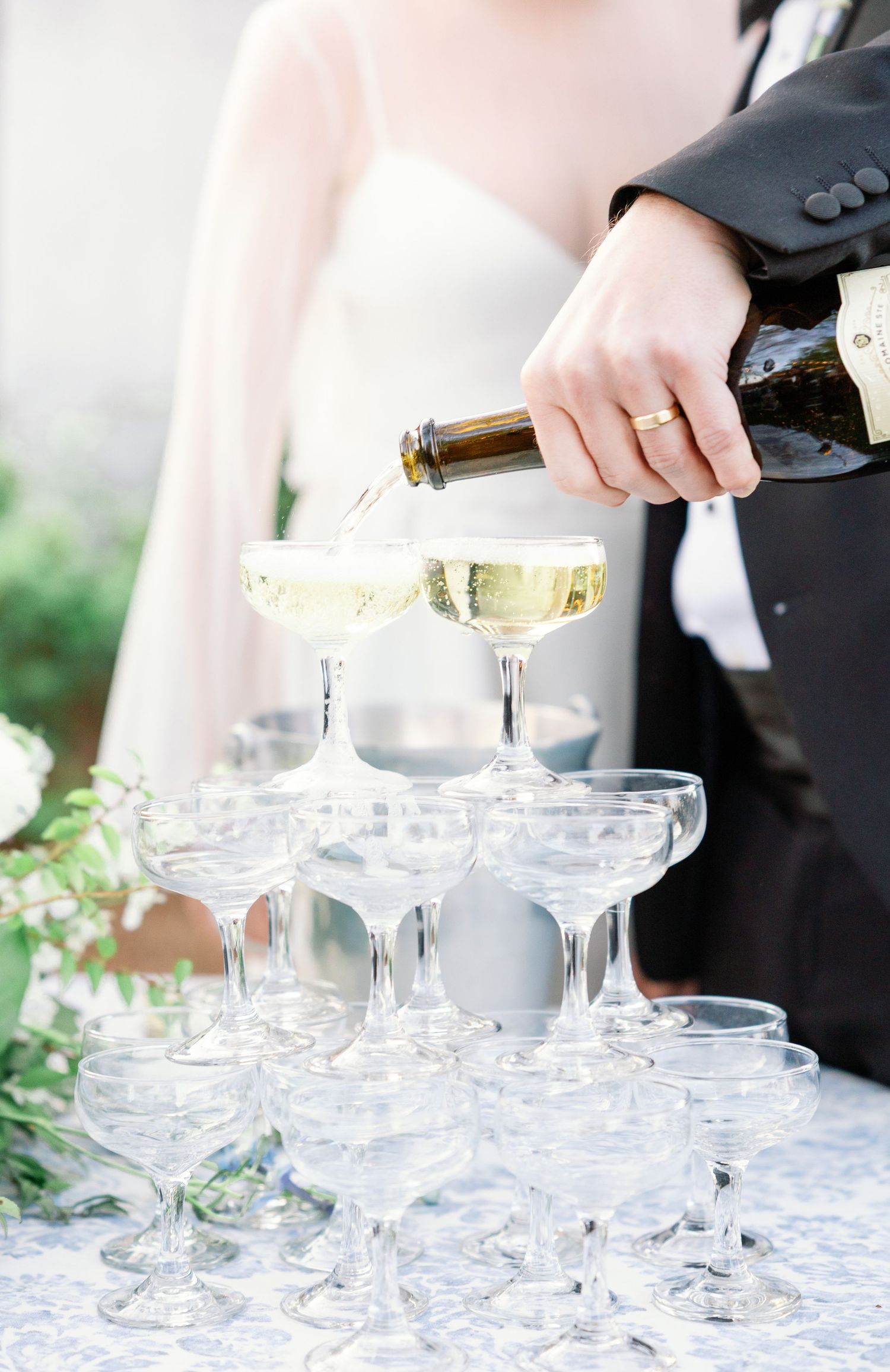 Champagne being poured into a tower of coupe glasses at an elegant event with delicate floral decorations.