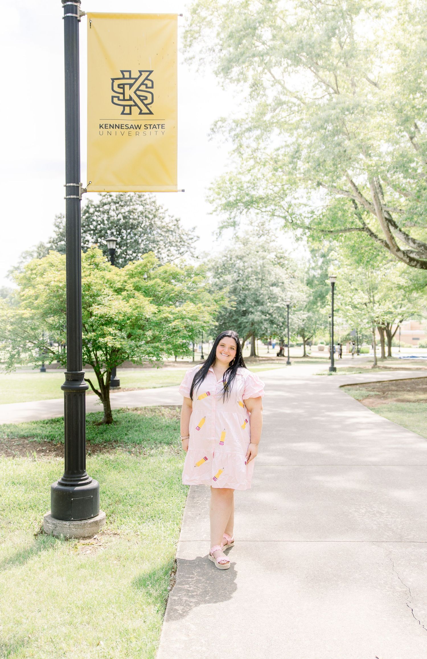 Graduate poses beneath yellow Kennesaw State University banner on campus path.