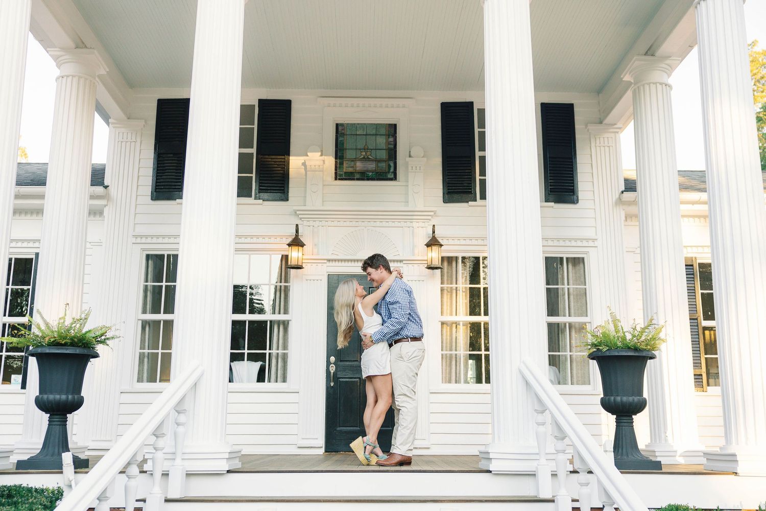 A couple embraces on the front steps of a white columned southern mansion with black shutters and potted plants.
