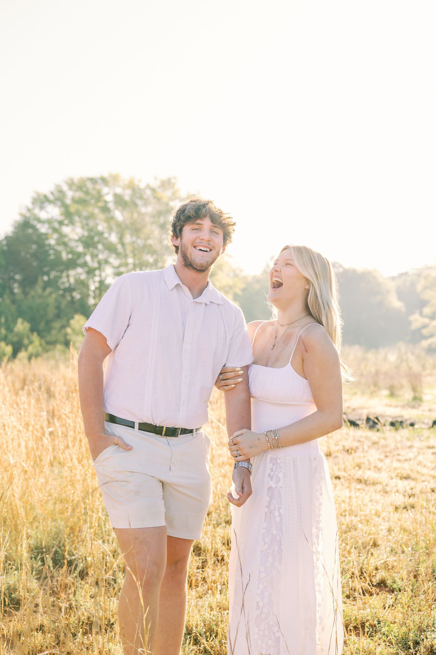 Joyful couple laughing together in golden sunlit field wearing white summer outfits.