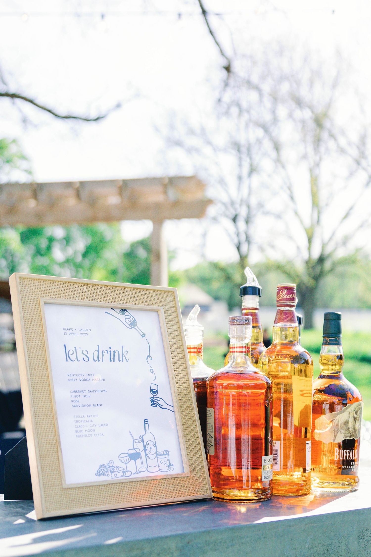 Bar setup with framed cocktail menu and assorted liquor bottles at outdoor wedding reception.