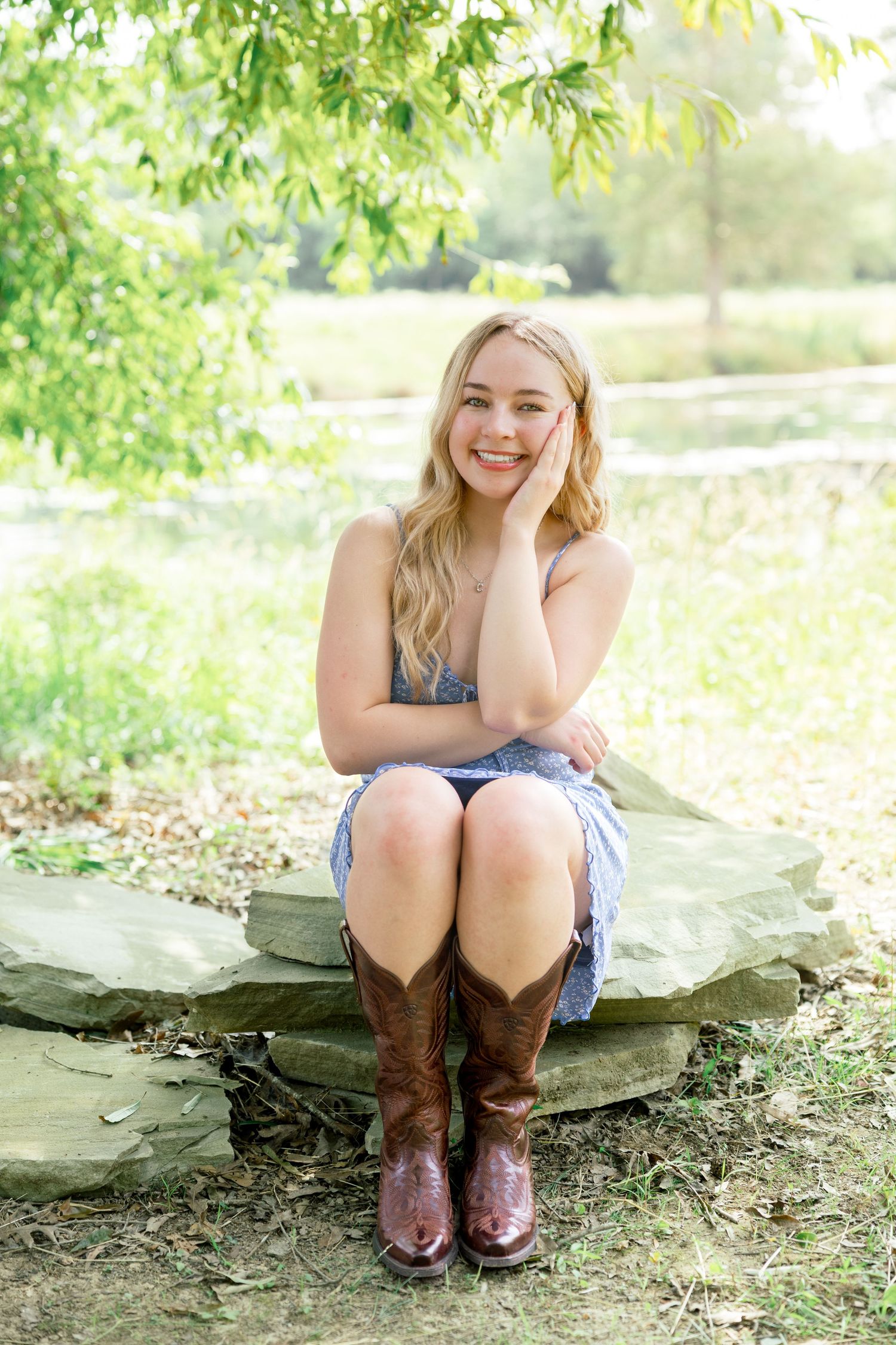 A person in a blue dress and brown cowboy boots sits on a large rock in a sunny outdoor setting with green foliage.