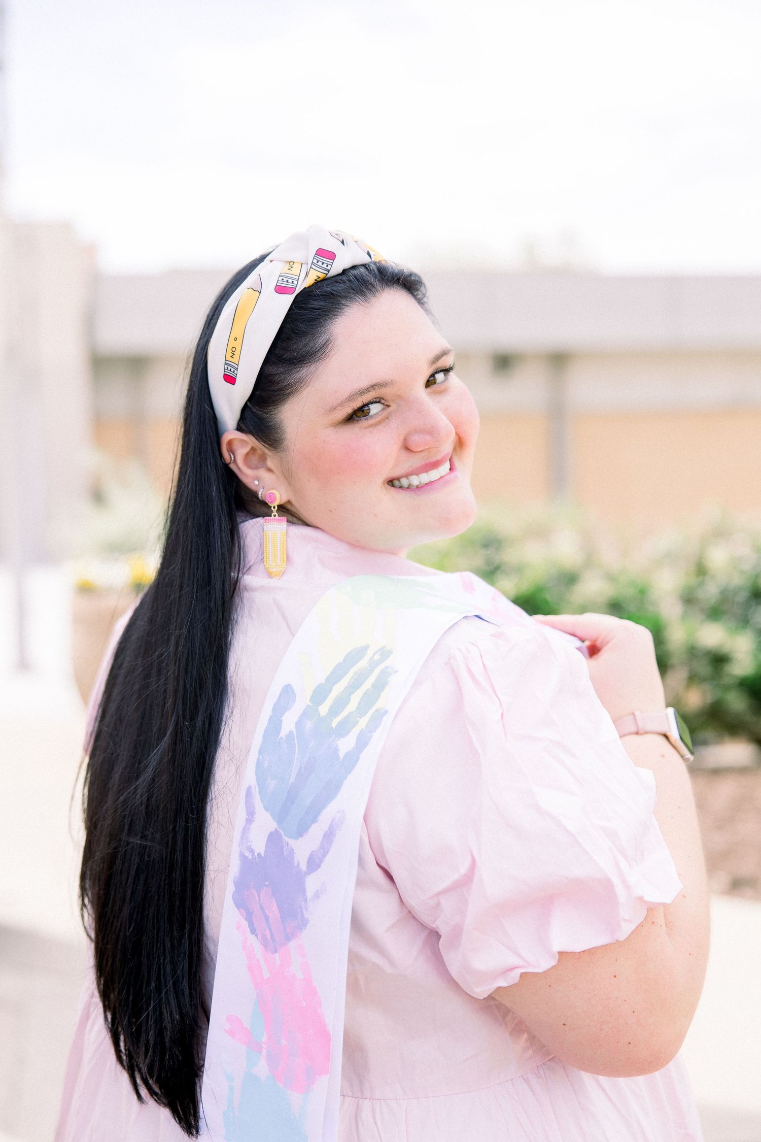 Person in pink dress with handprint sash and pencil headband smiling outdoors.