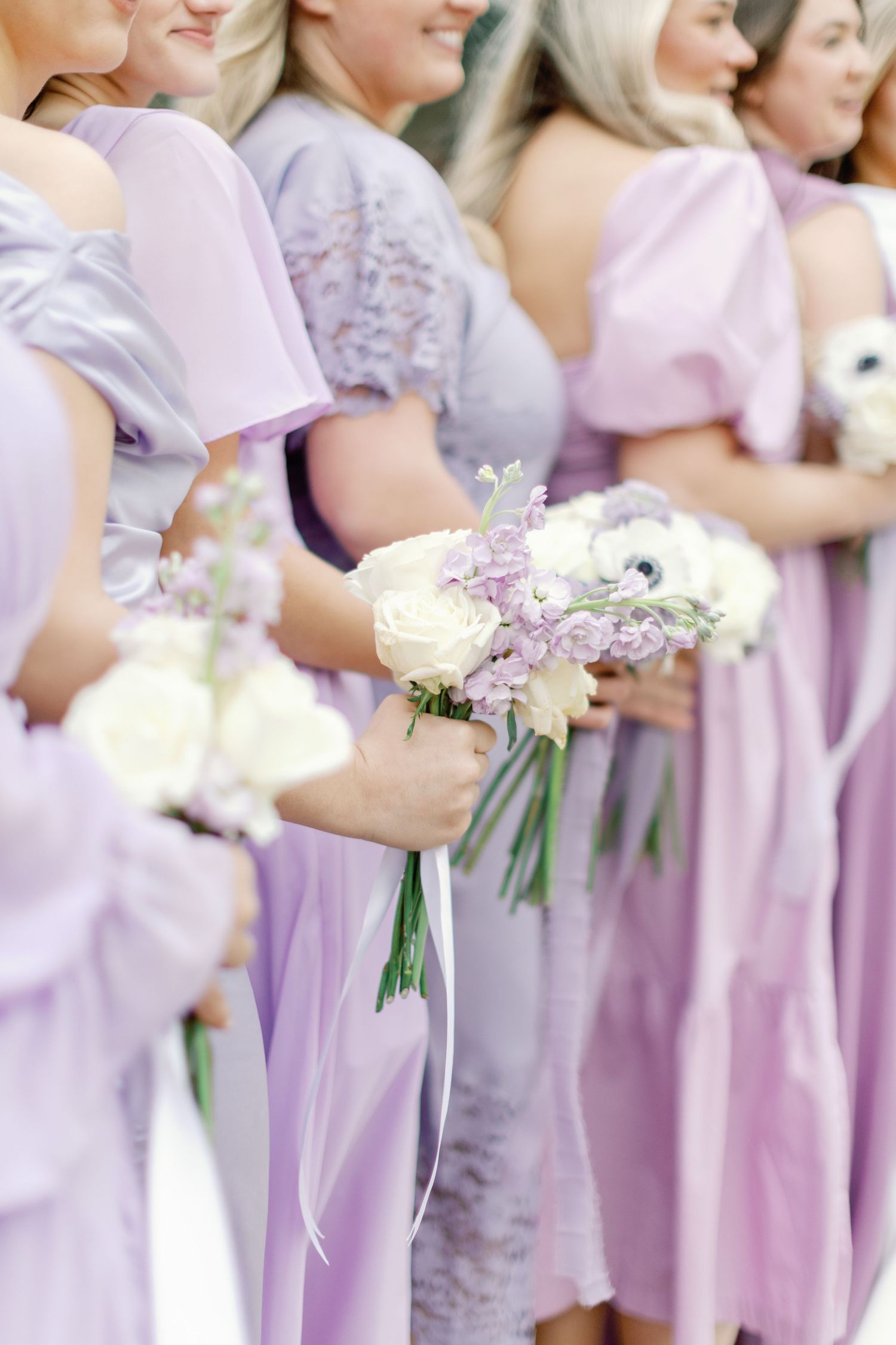 A row of bridesmaids in soft lilac dresses holding delicate white and purple flower bouquets at a wedding ceremony.