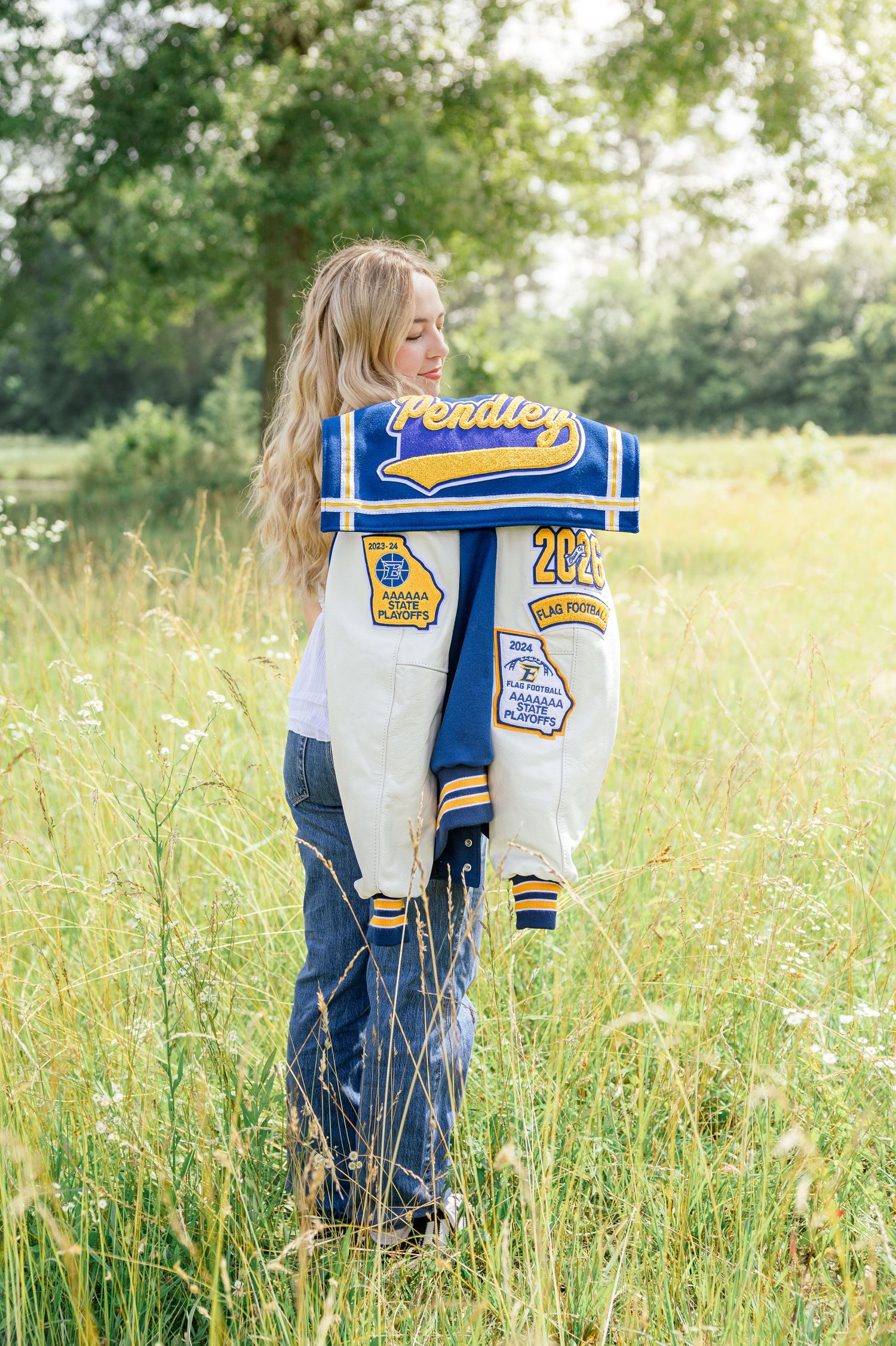 Student in jeans holds a blue and white varsity letterman jacket with patches in a sunny meadow.