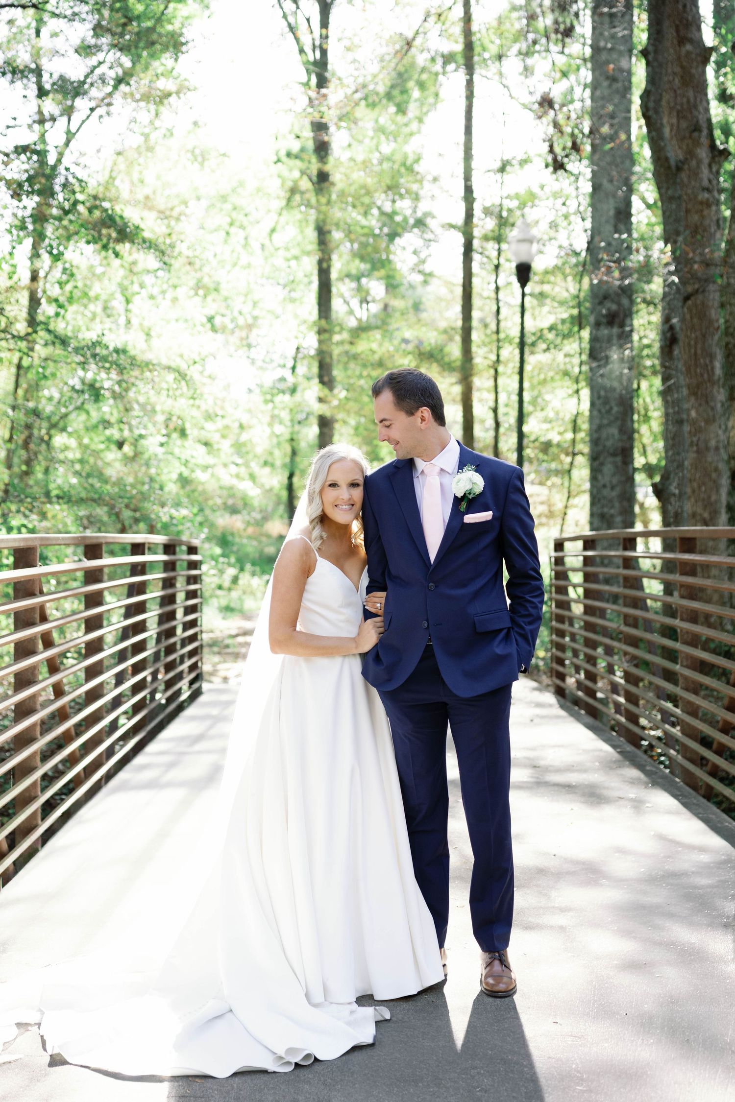 A couple stands together on a wooden footbridge surrounded by lush green forest on their wedding day.