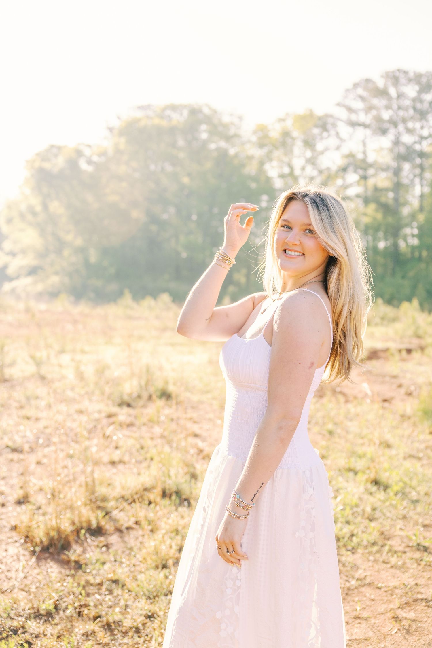 A woman in a white sundress poses gracefully in a sunlit field during golden hour.