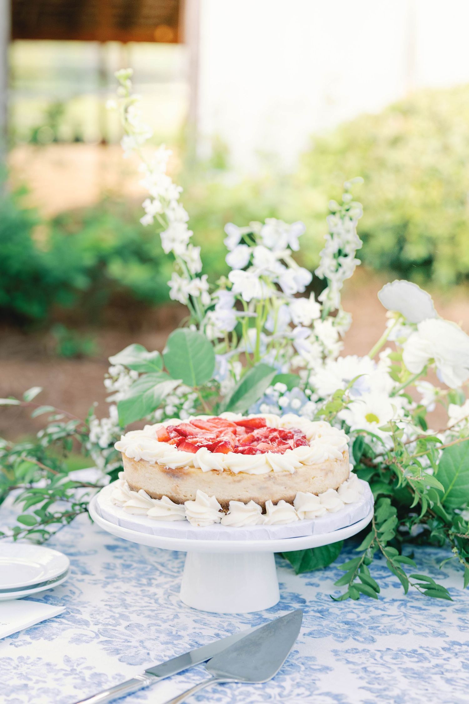 White frosted cake with strawberry topping displayed on blue patterned tablecloth with floral decor.