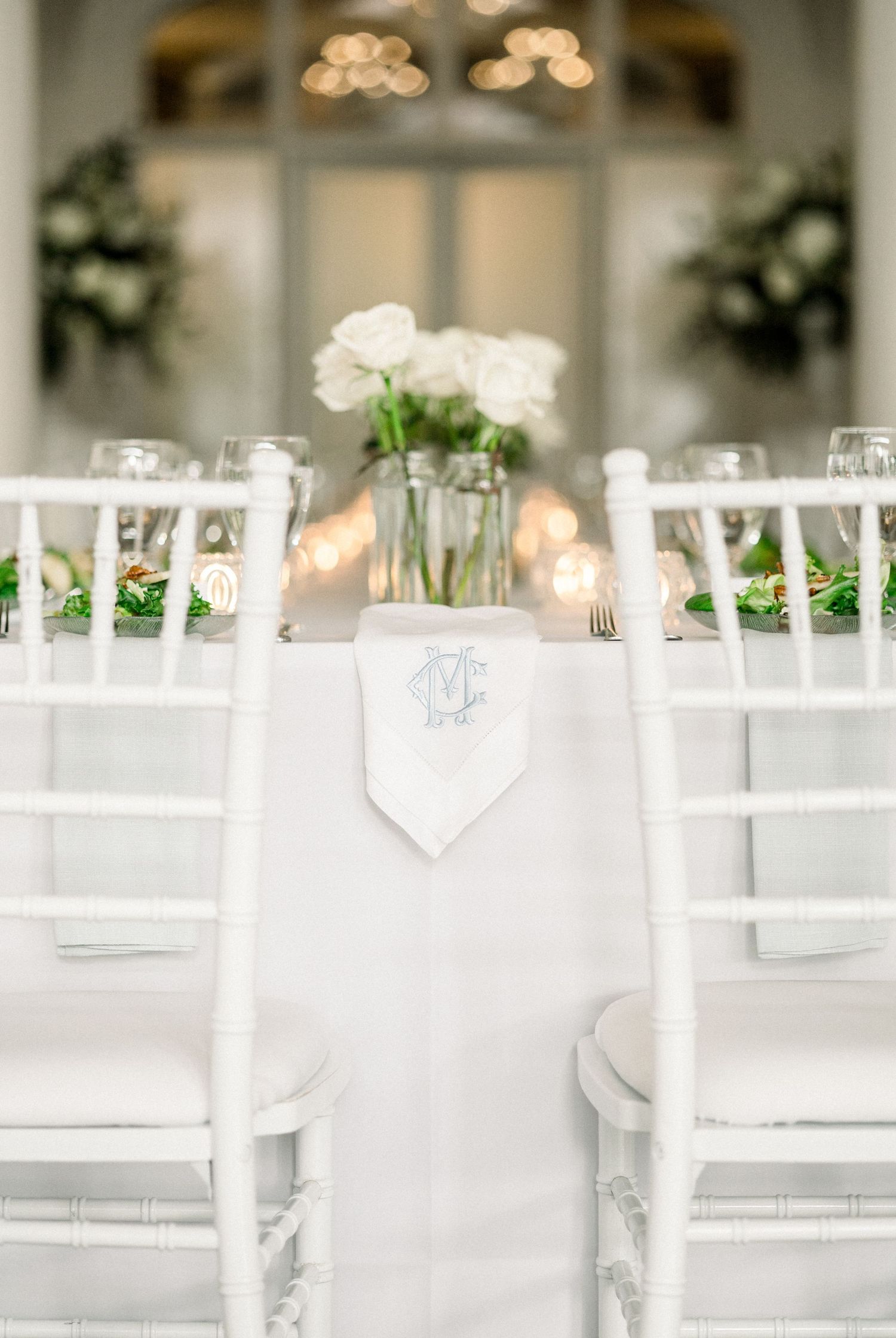 Elegant white Chiavari chairs and monogrammed napkins set for an upscale wedding reception with floral centerpieces.