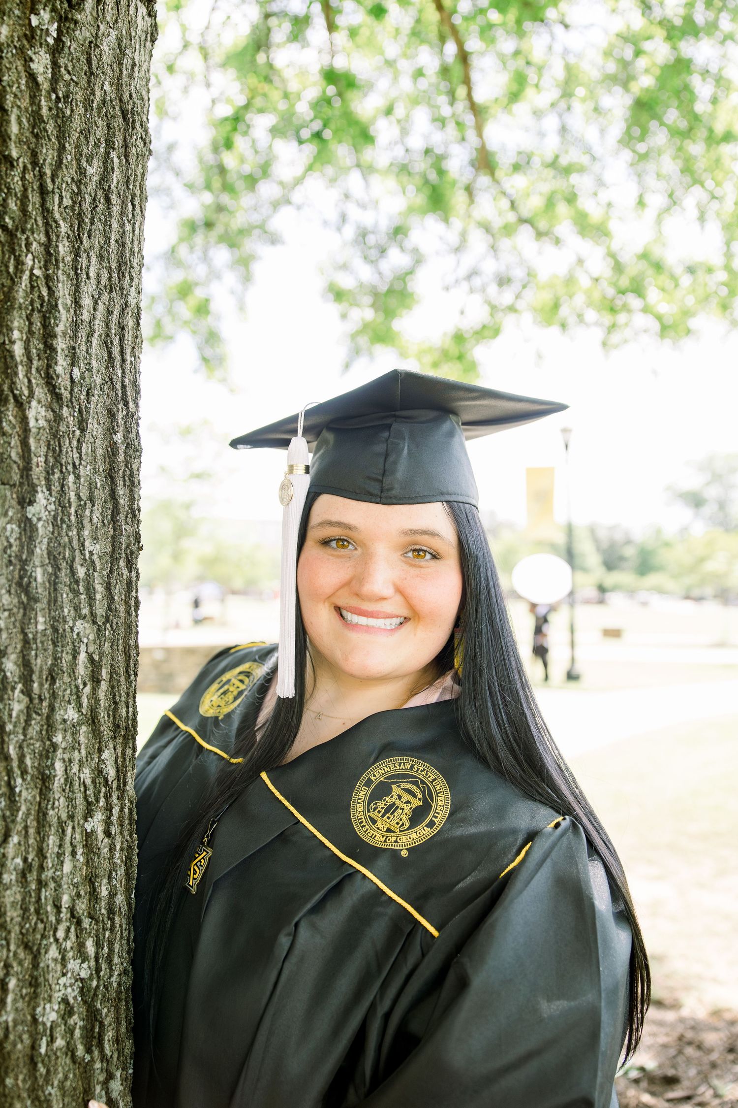 Graduate in black cap and gown with honors stole standing next to tree.