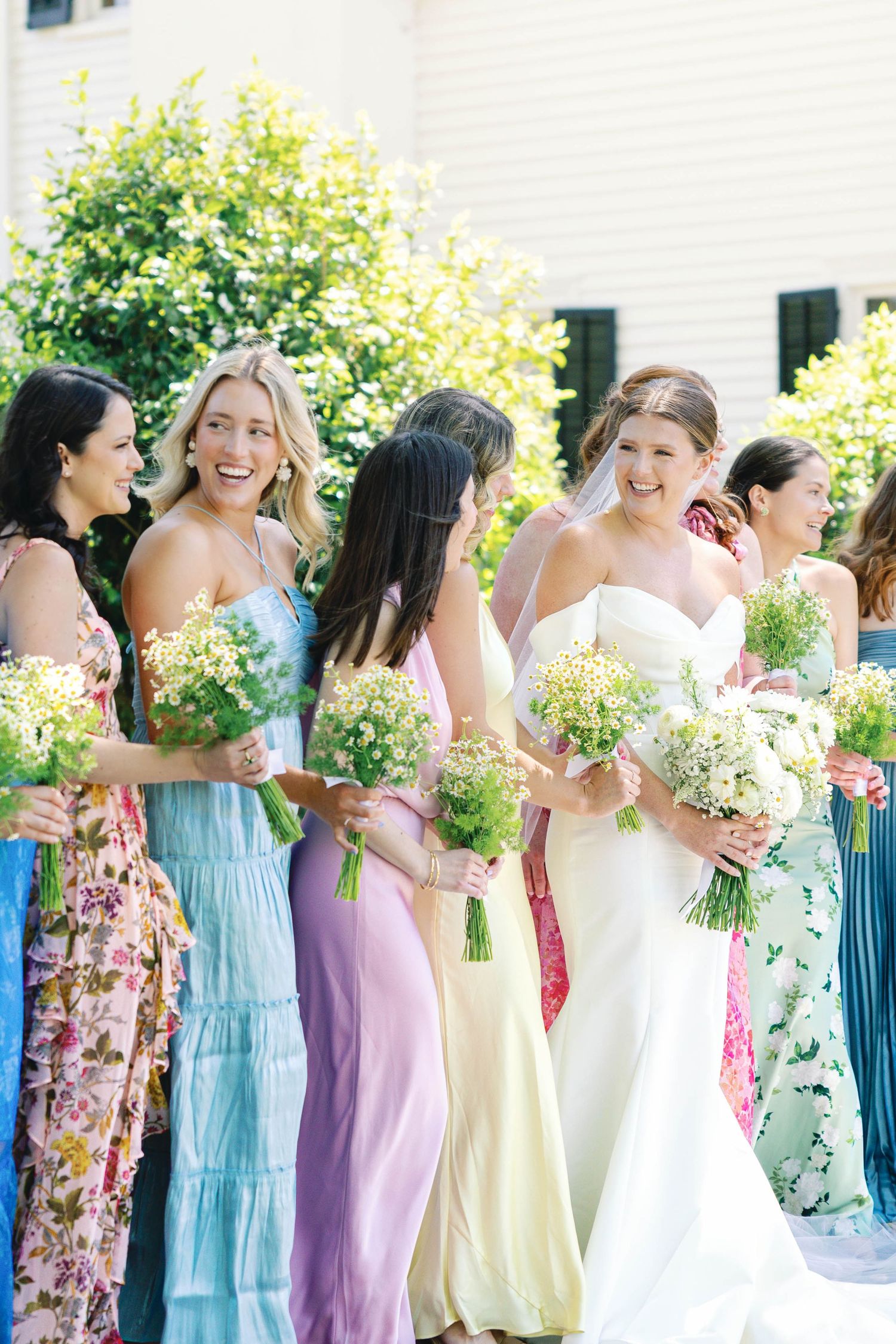 A line of bridesmaids in pastel floral and solid dresses stand together holding bouquets at an outdoor wedding.