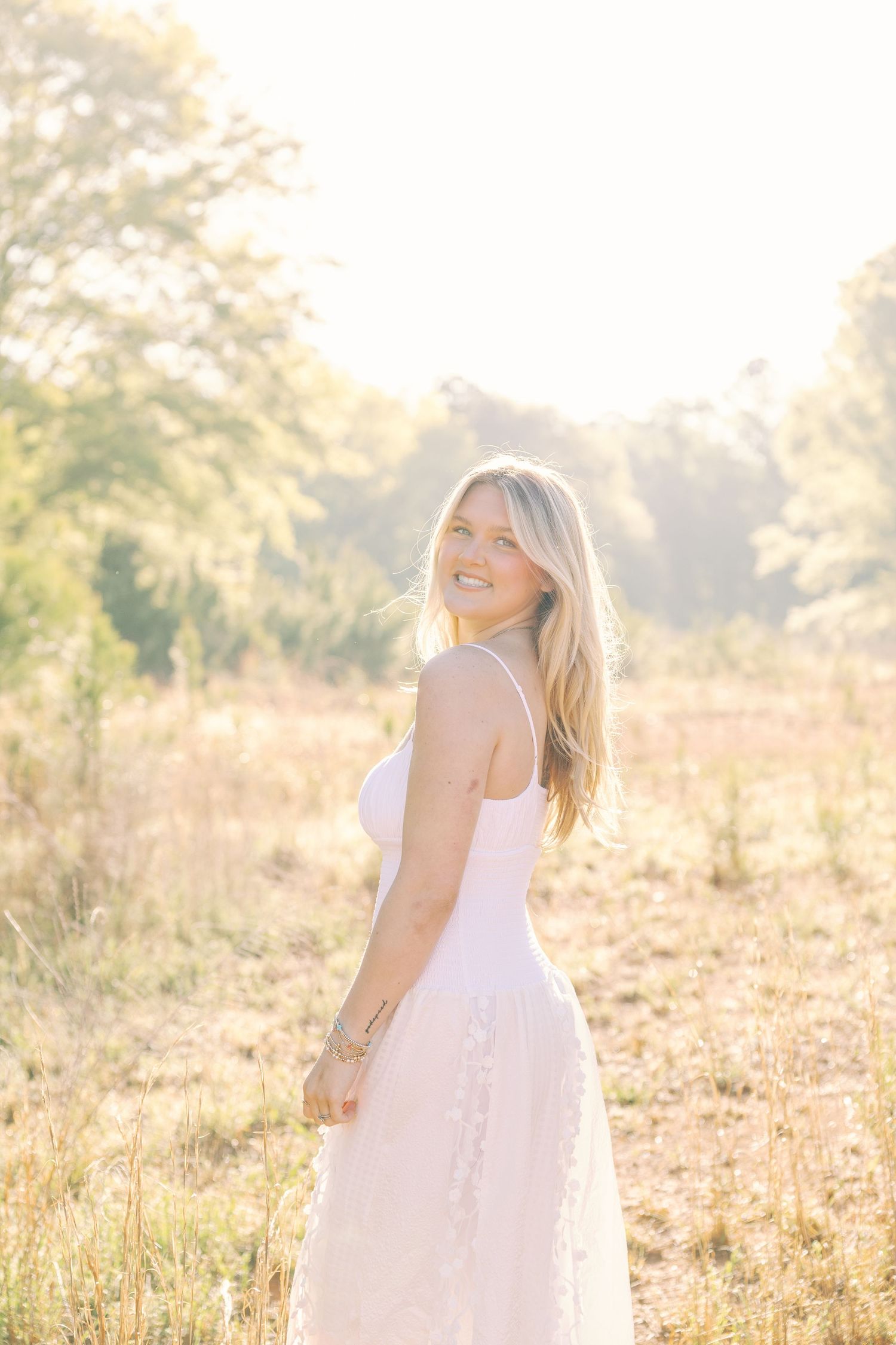 Woman in white sundress smiles while posing in a sun-drenched summer field.