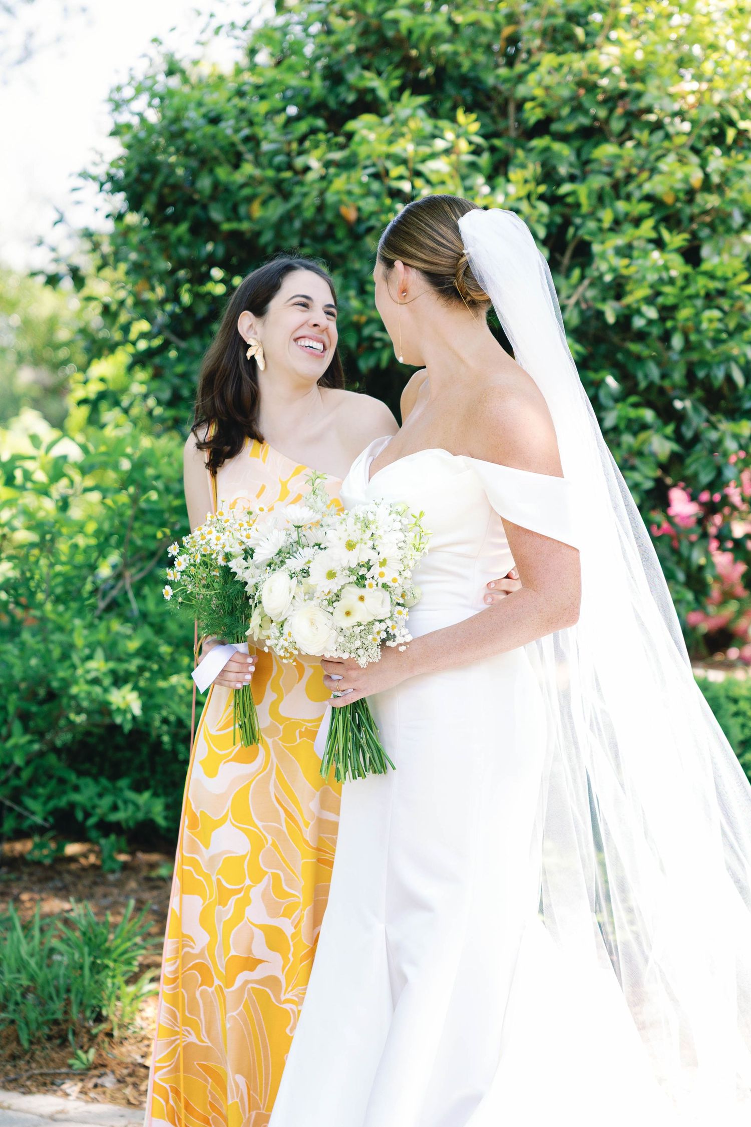 Two wedding attendees share joyful moment in garden setting with floral bouquets.