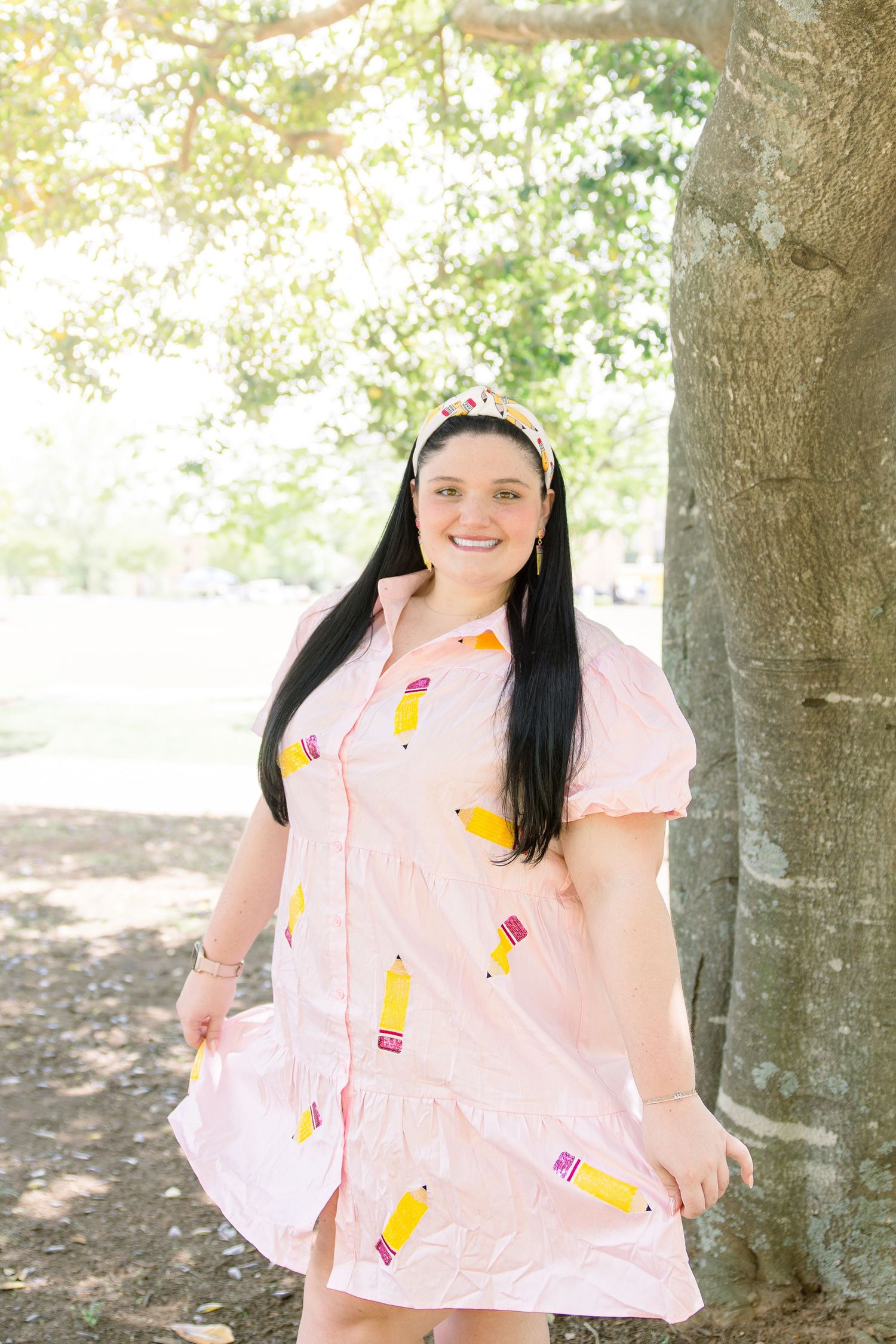 Person in light pink dress with pencil pattern standing by tree trunk outdoors.