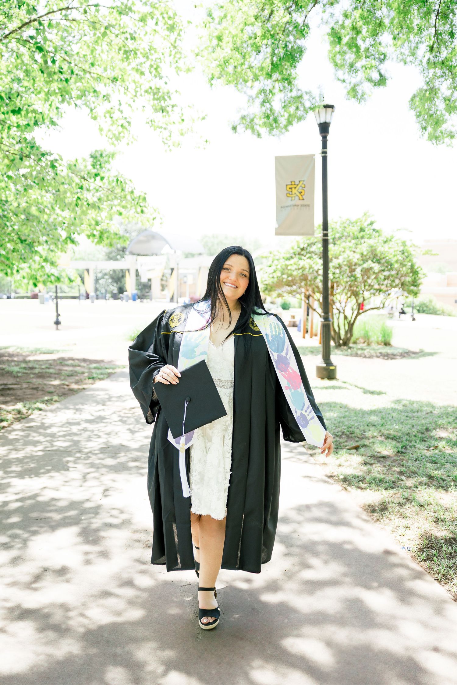 Graduate in black cap and gown walking on campus path holding diploma.