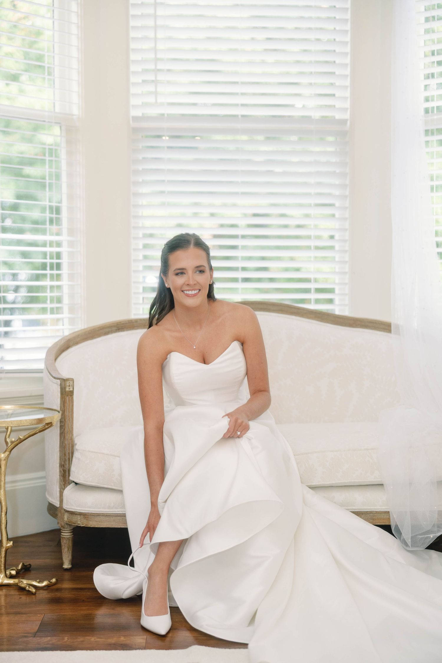 Elegant bridal portraits showing a white wedding gown on a cream-colored French settee by window light.