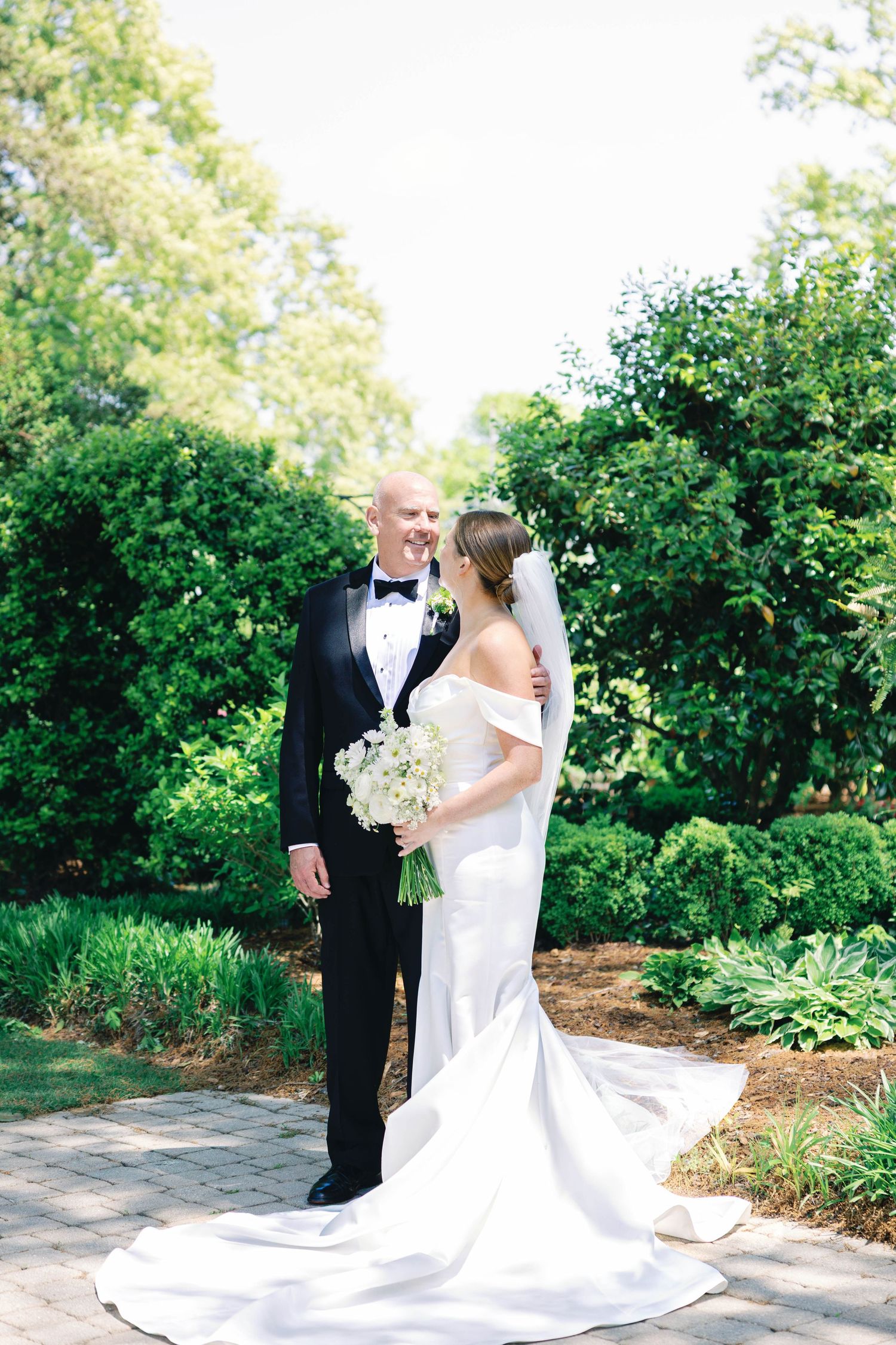 Newlyweds pose in garden setting with bride wearing flowing off-shoulder gown with long train.