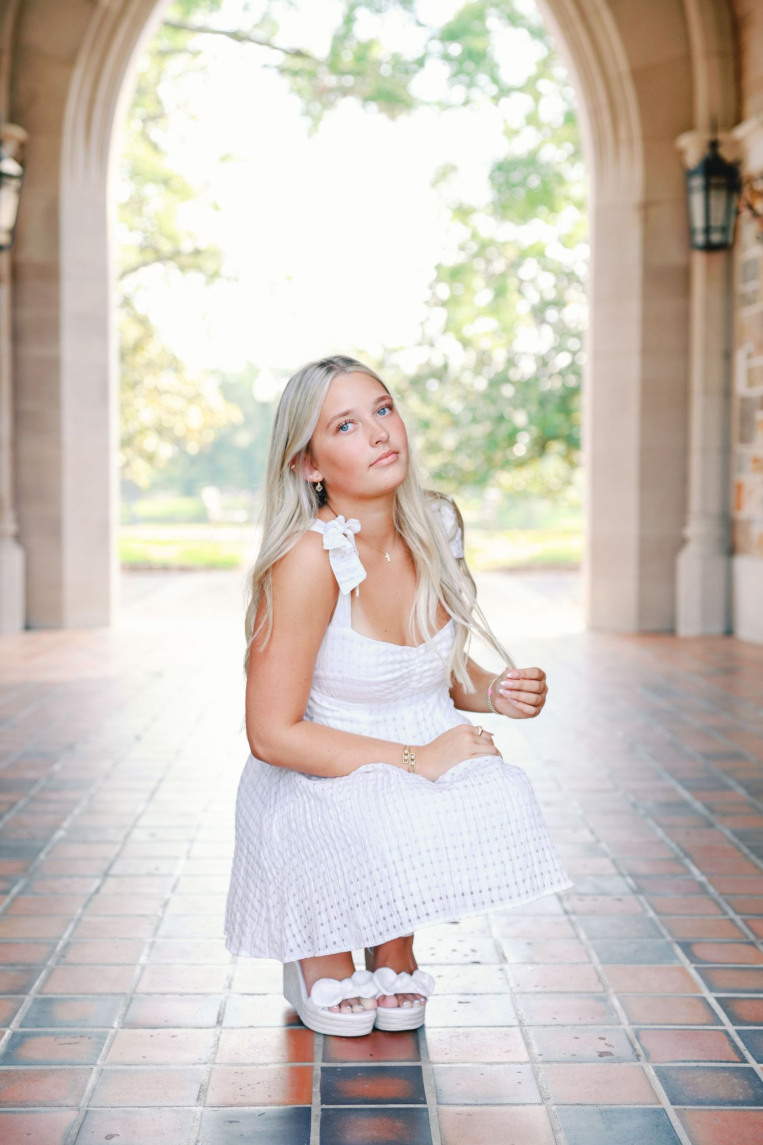 A person in a white eyelet lace dress poses on tiled ground under an arched architectural walkway with sunlit trees behind.