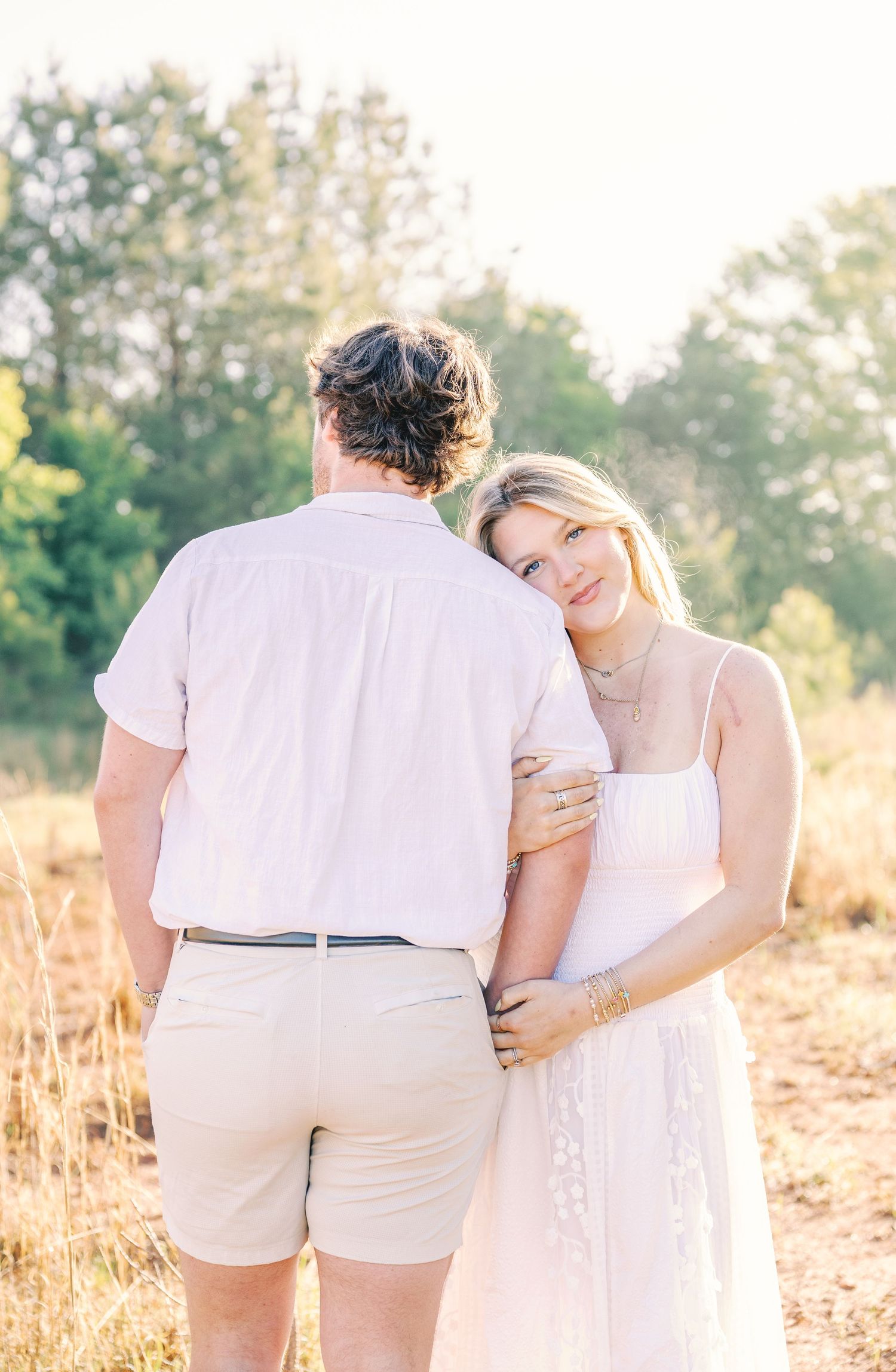 Couple in white summer clothing embracing in golden sunlit field during sunset.