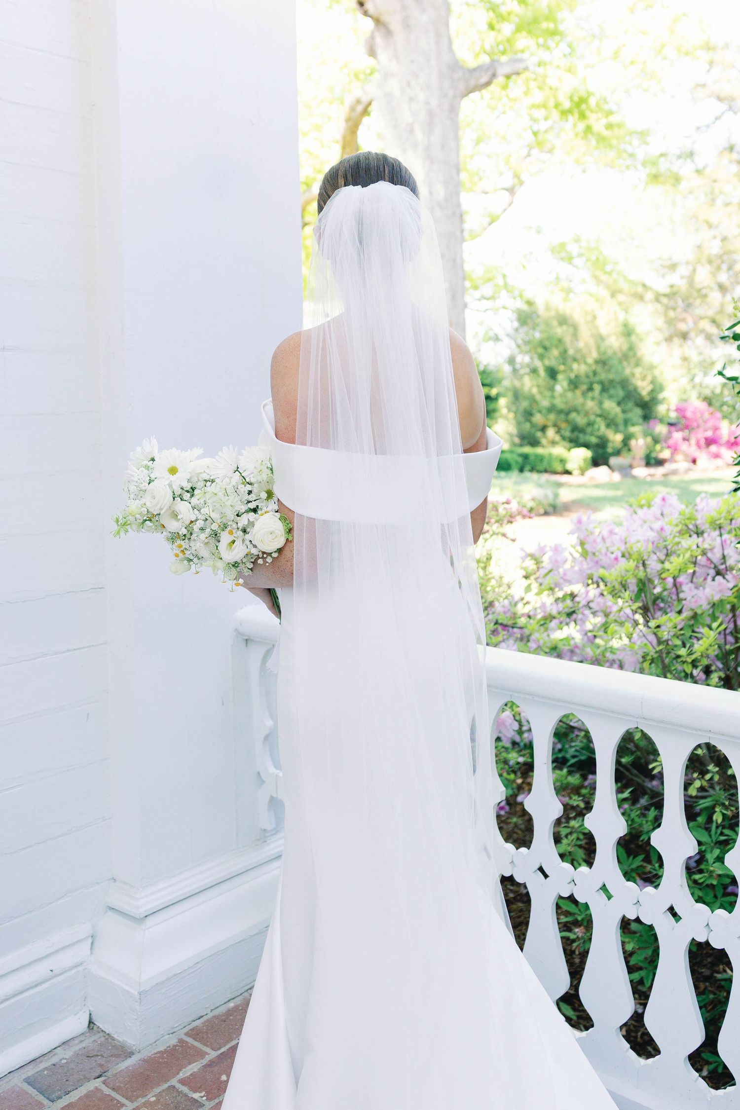 Back view of bride in fitted white gown and veil standing on columned porch.