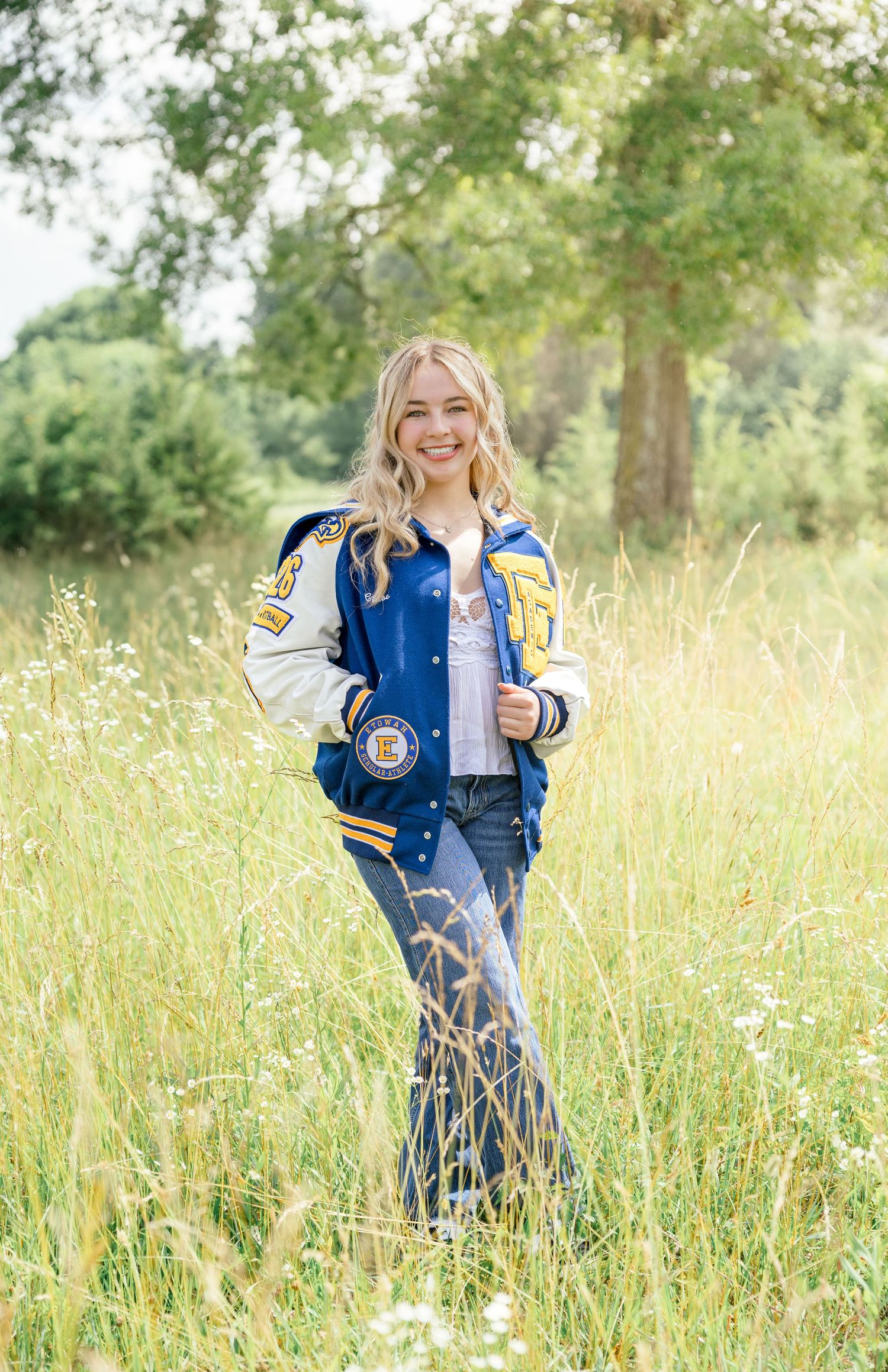 A person wearing a blue and gold varsity letterman jacket poses in a sunlit field of tall grass and wildflowers.