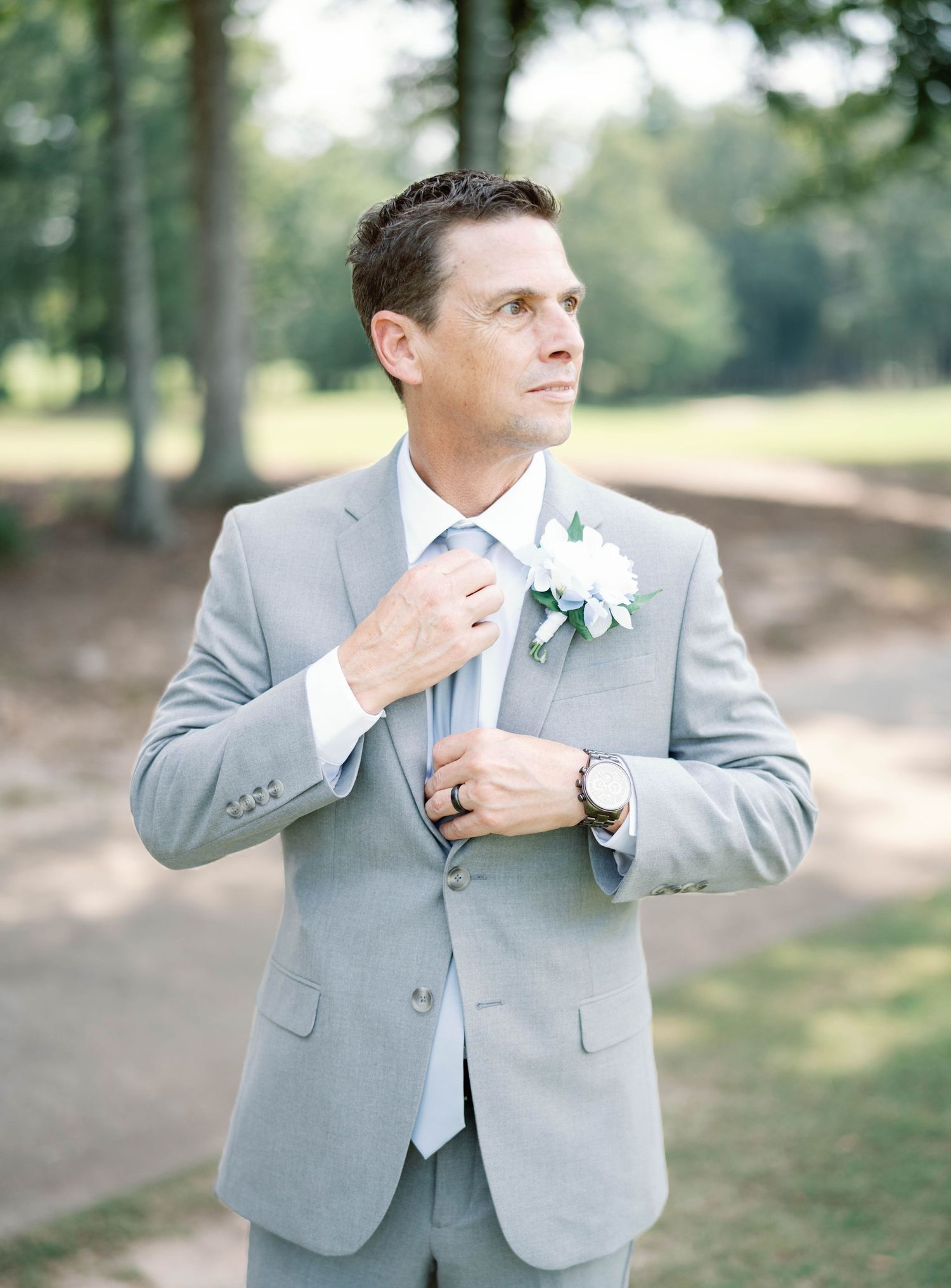 A person wearing a light gray suit jacket adjusts their tie while standing outdoors in a natural setting.