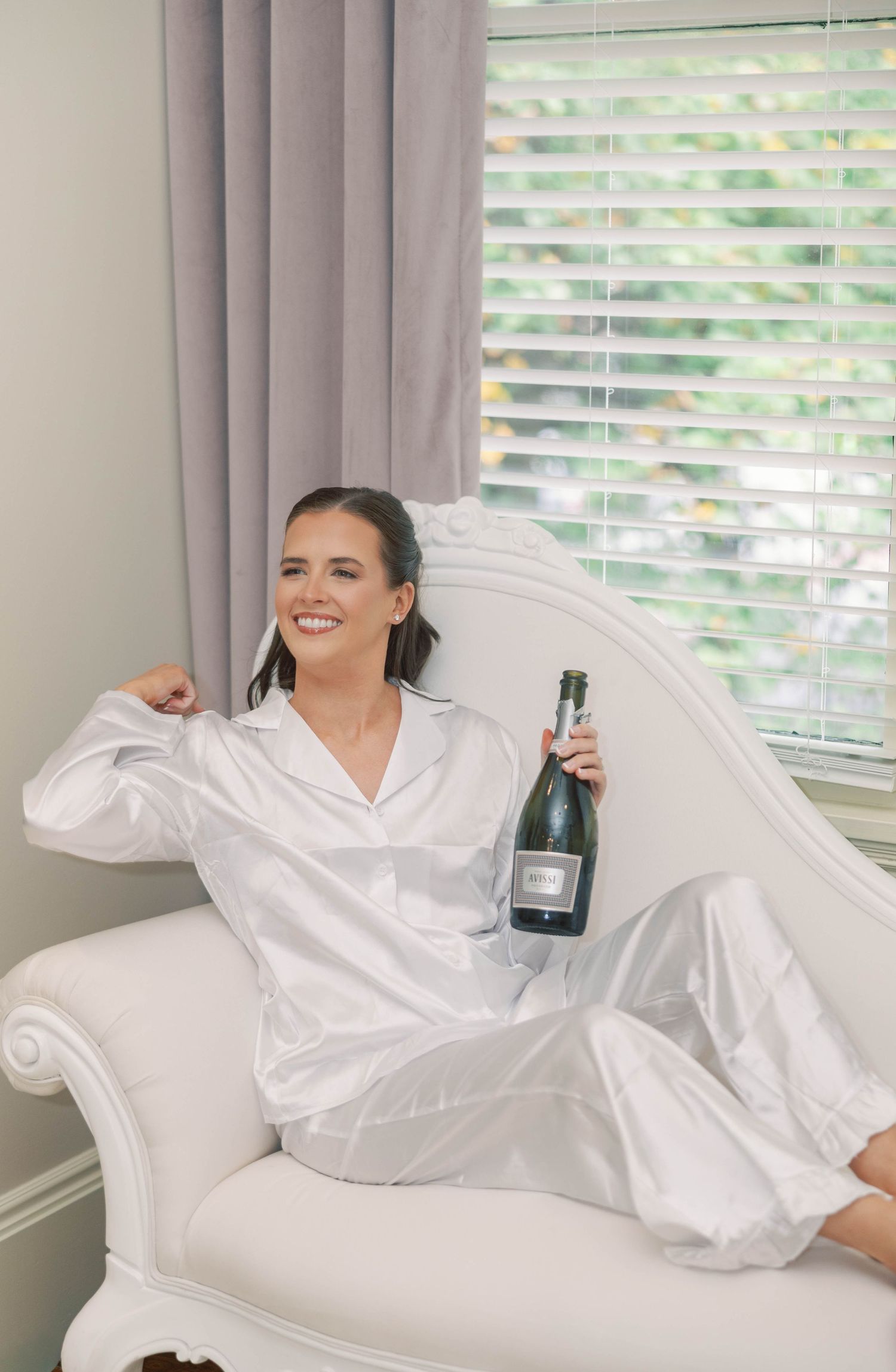 Person in white robe lounges on white couch with champagne bottle in bright sunlit room.
