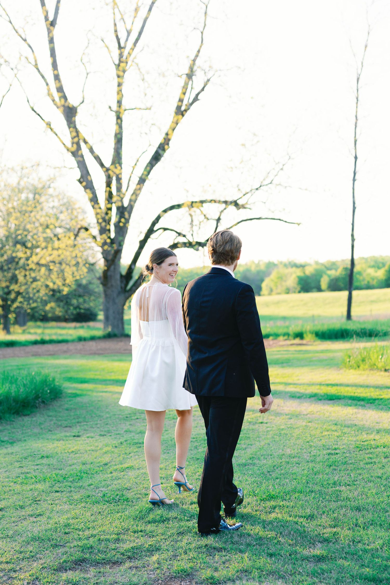 A couple in formal attire walks through a scenic park setting during golden hour.