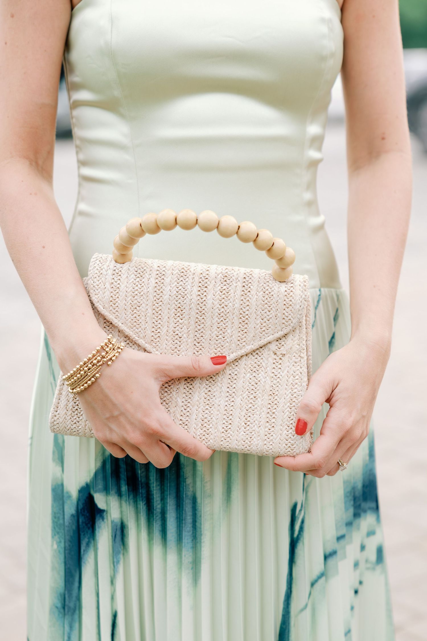 Woman in mint dress holding a woven cream clutch bag with wooden bead handle and red manicured nails.