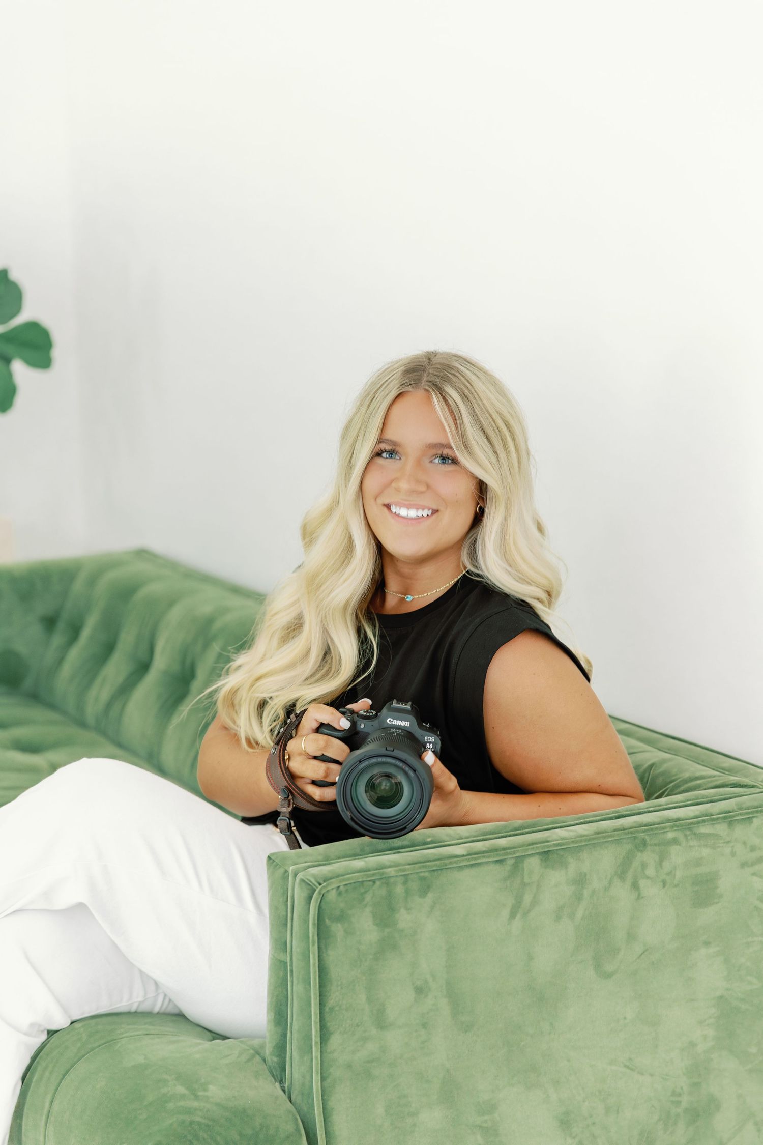 Professional photographer with blonde hair lies on green velvet couch holding camera in casual lifestyle photoshoot setting.