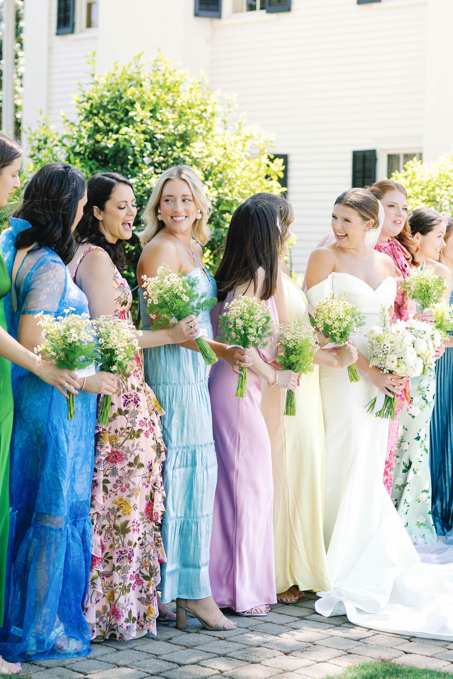 Bridesmaids in pastel and floral dresses holding bouquets while standing with bride on brick patio.