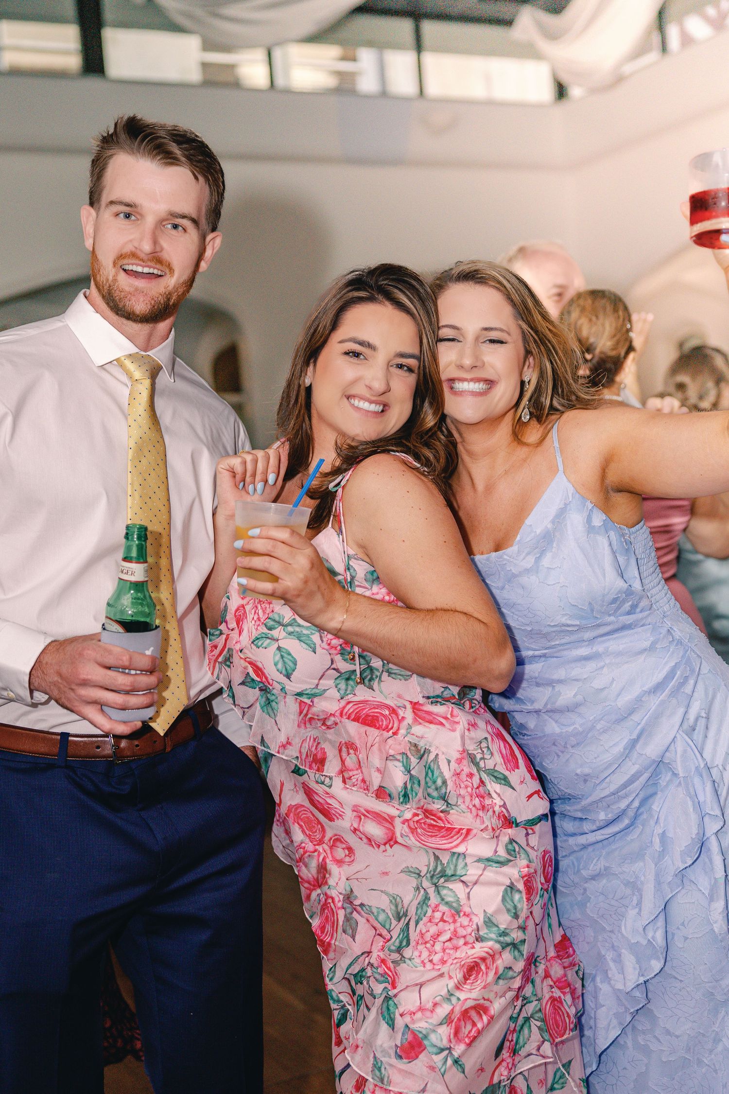 Three friends enjoying drinks and celebrating at a festive indoor party, wearing formal attire in pastel and floral patterns.