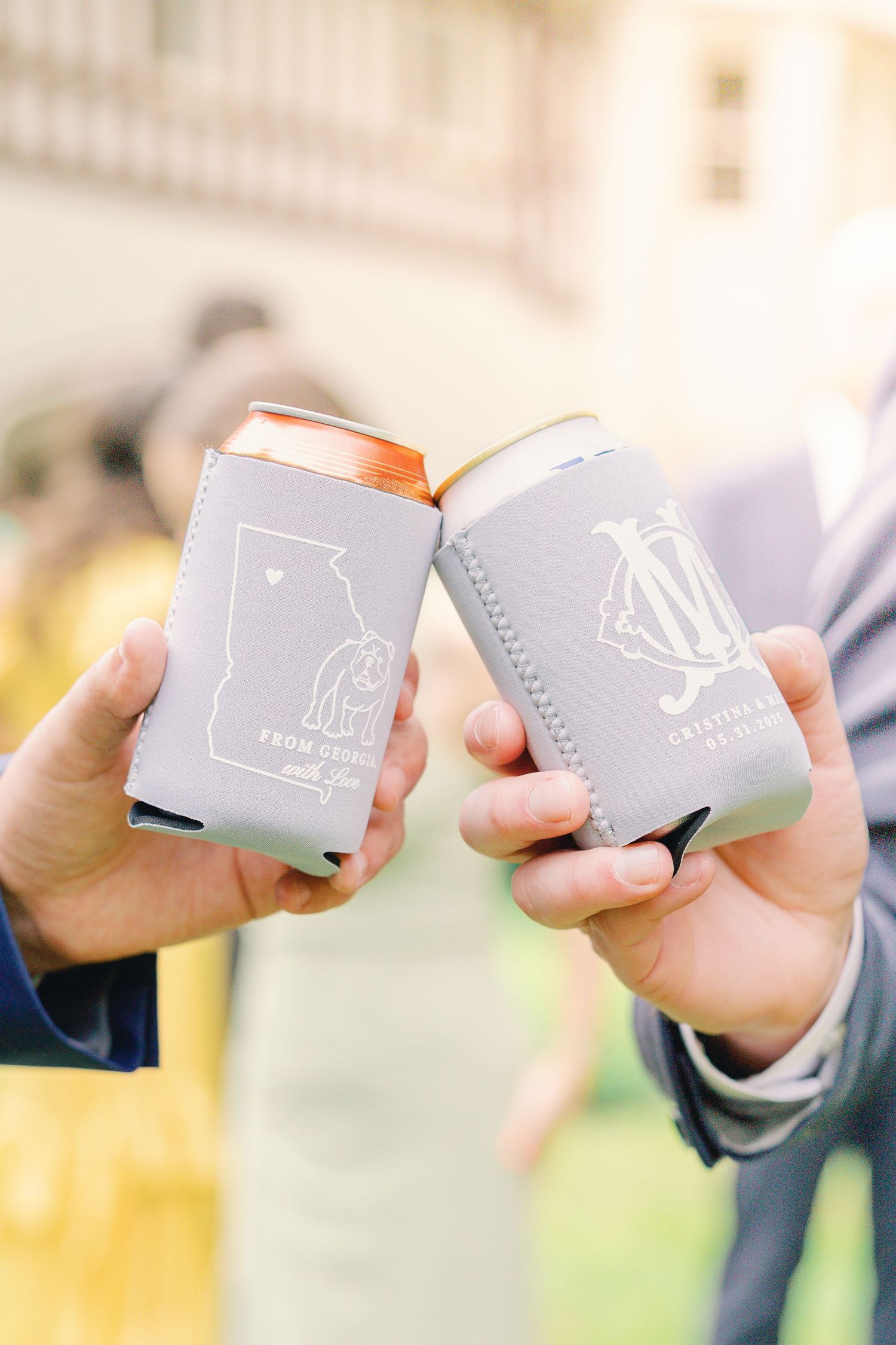 Two hands clink beverage cans with gray koozies featuring custom monogram designs in an outdoor setting.
