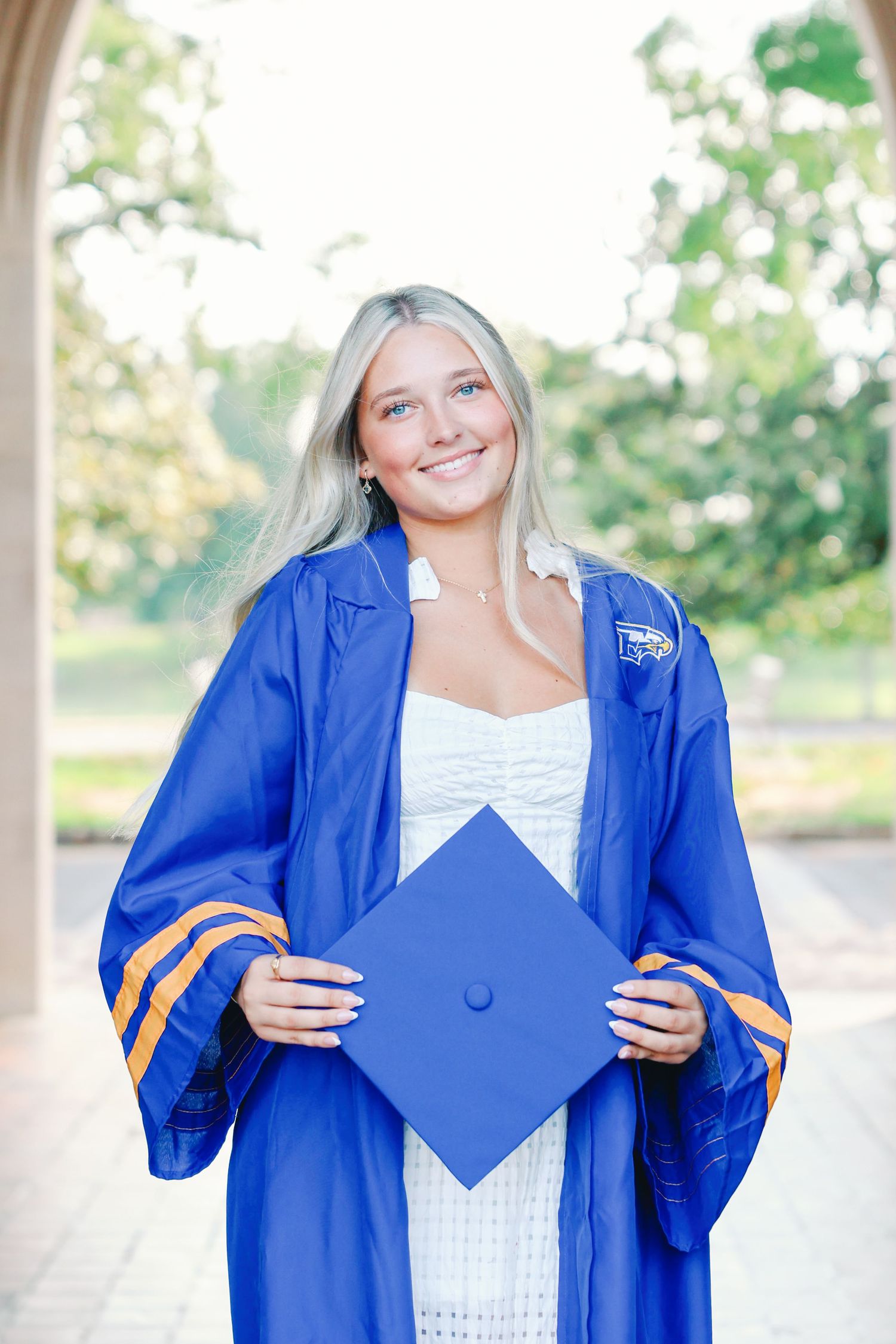 Graduate posing in bright royal blue cap and gown holding a graduation cap in front of a garden setting.