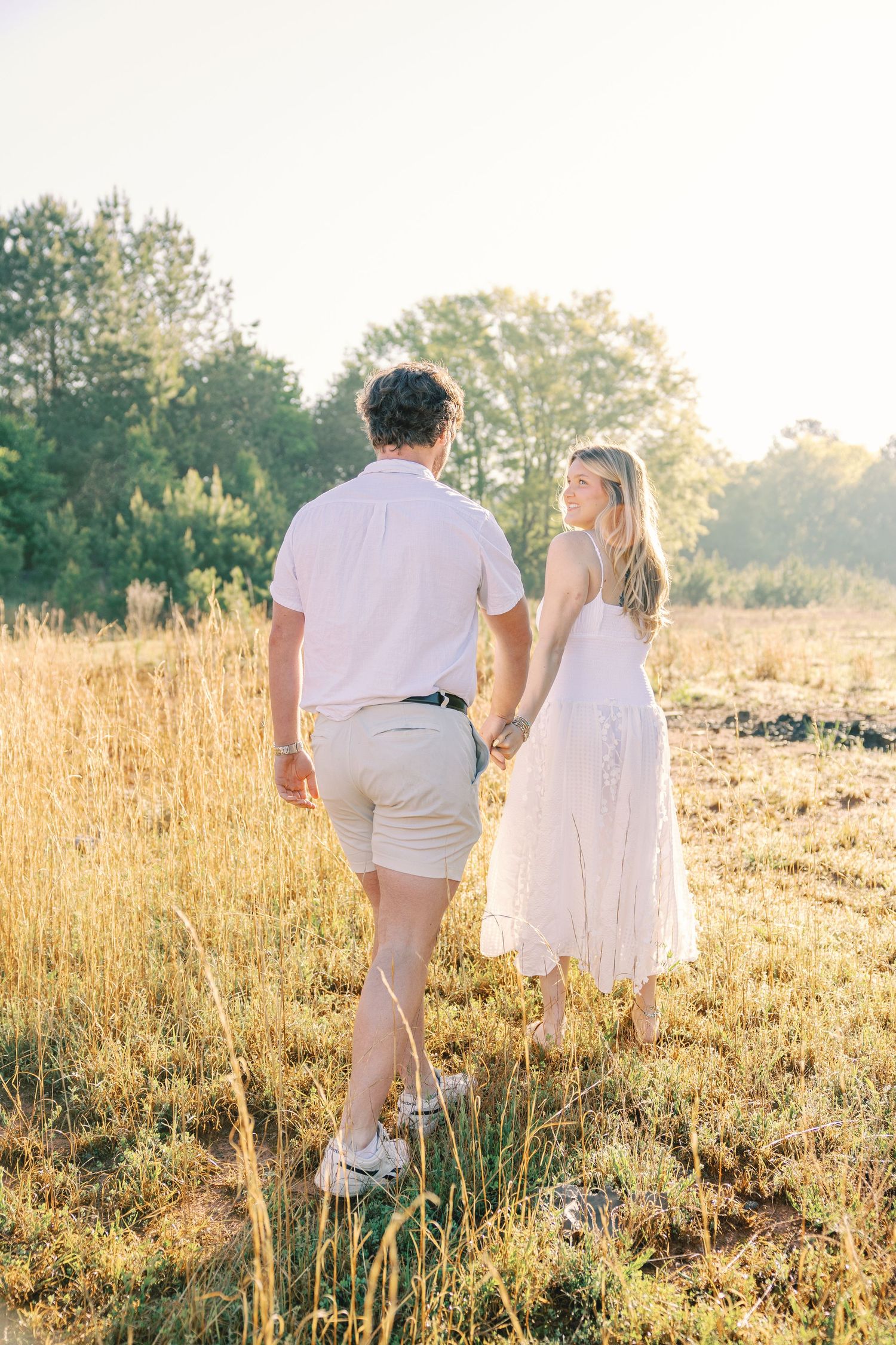 Couple walking hand in hand through sun-drenched field in white summer outfits.