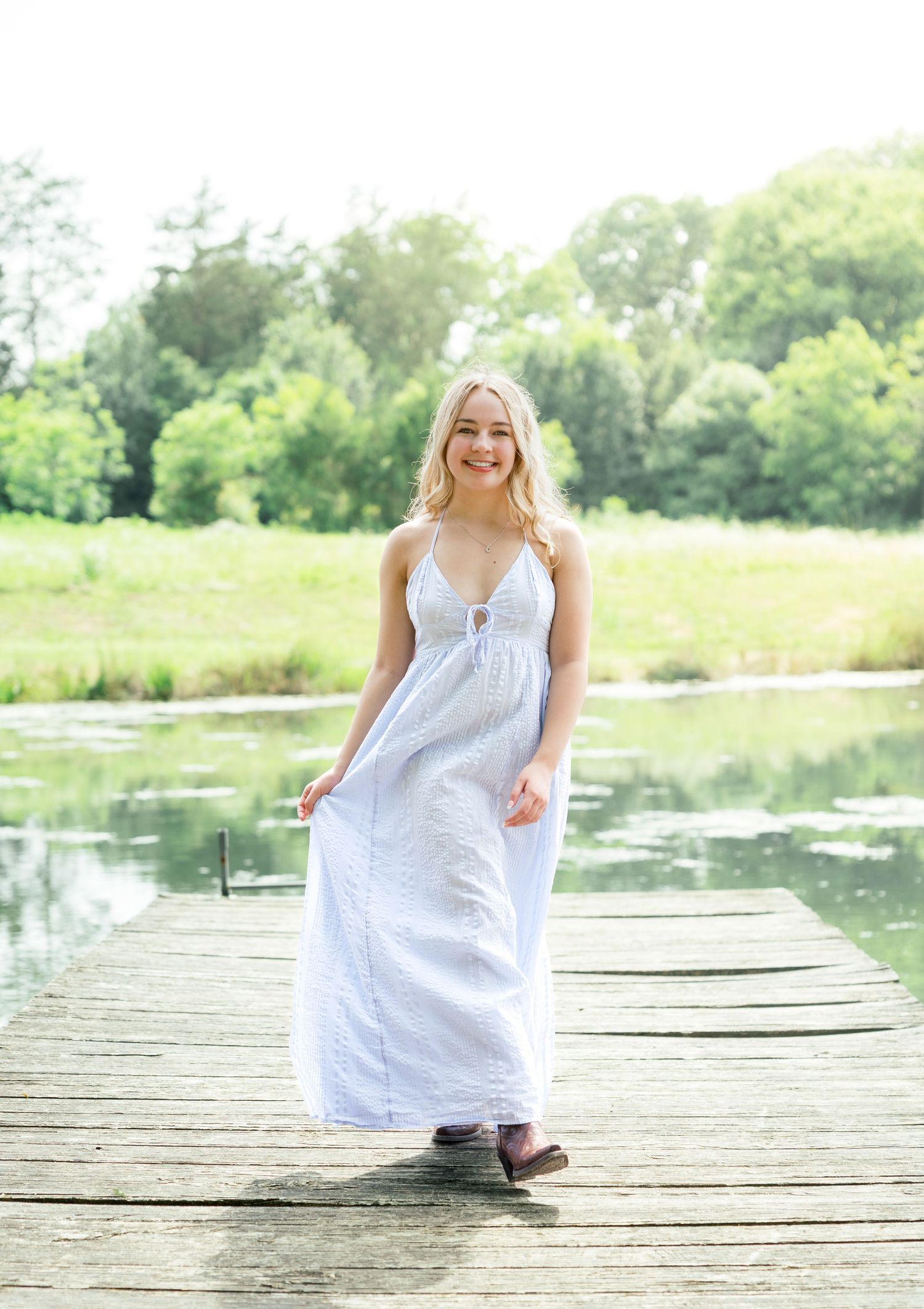 Person in a flowing white dress sits on a wooden dock by a serene lake surrounded by summer greenery.