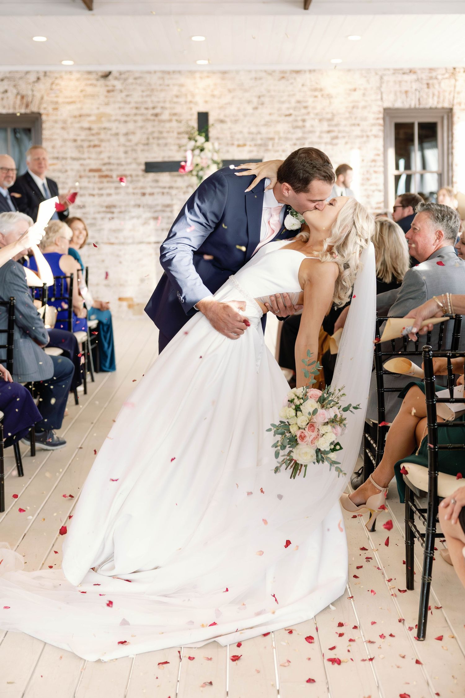 Romantic sequence of wedding couple sharing a dip kiss with rose petals falling in a rustic brick venue.