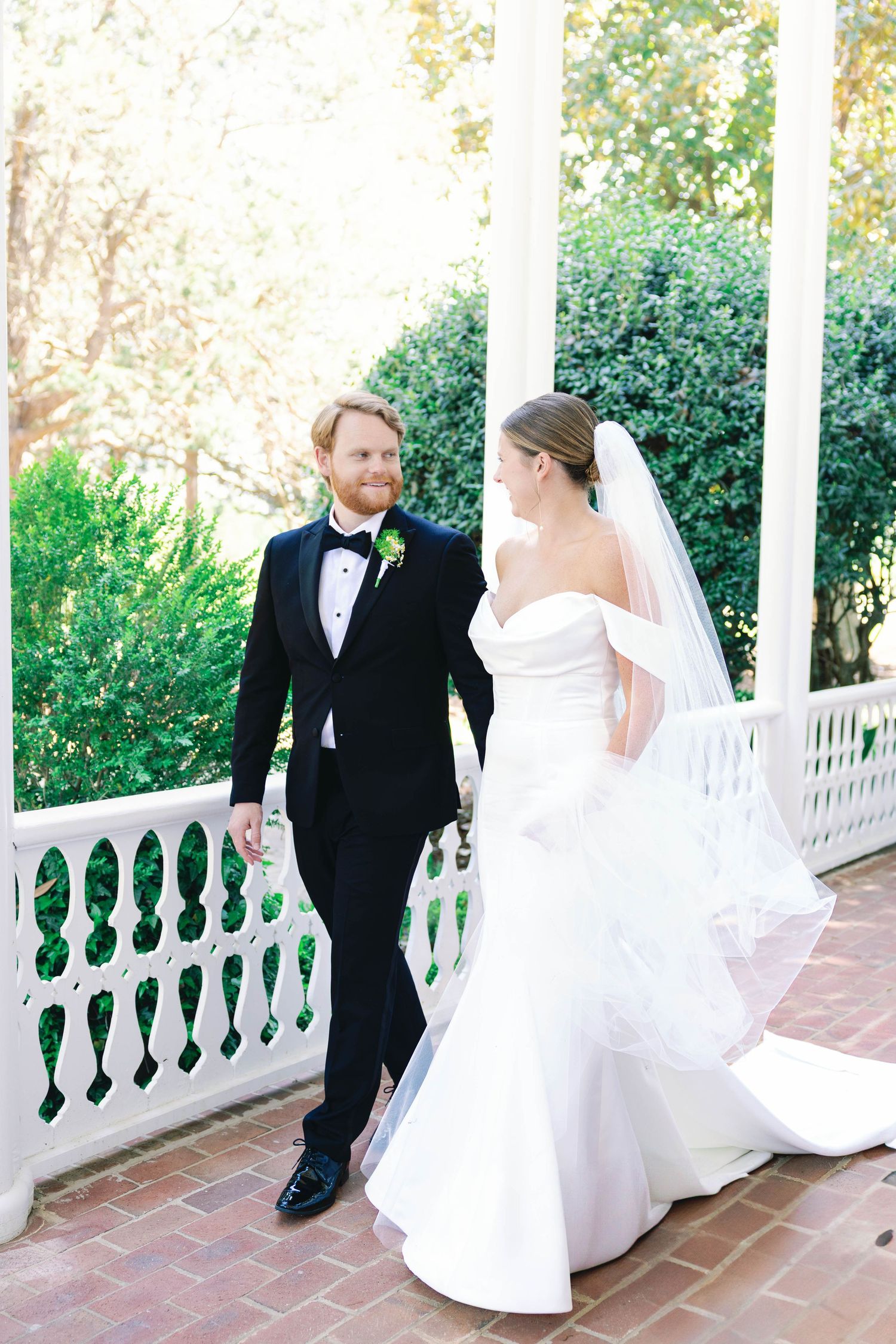 A couple in wedding attire stands on a classic white columned porch surrounded by greenery.