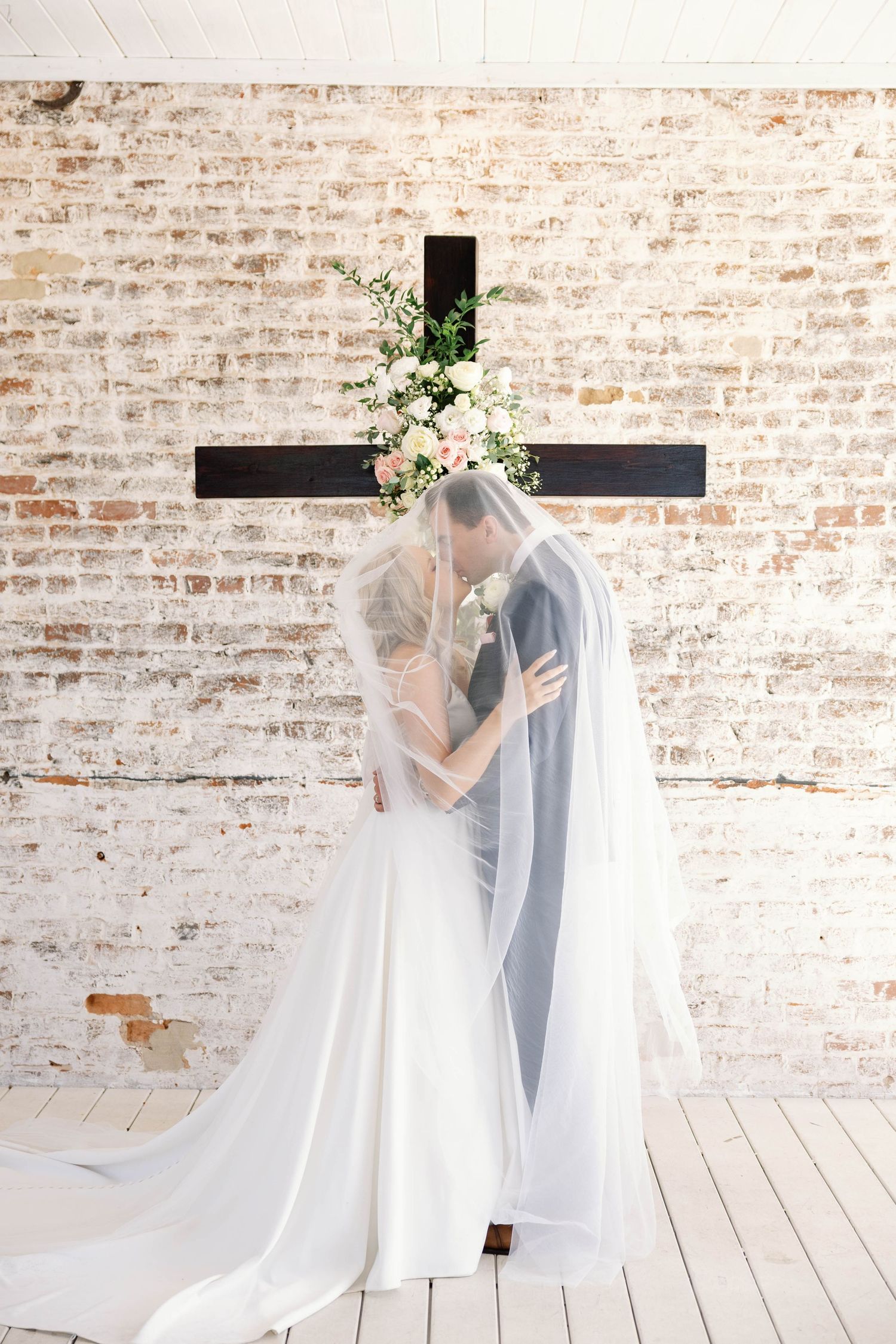 A romantic wedding scene showing a couple embracing under a delicate veil against a rustic exposed brick wall.