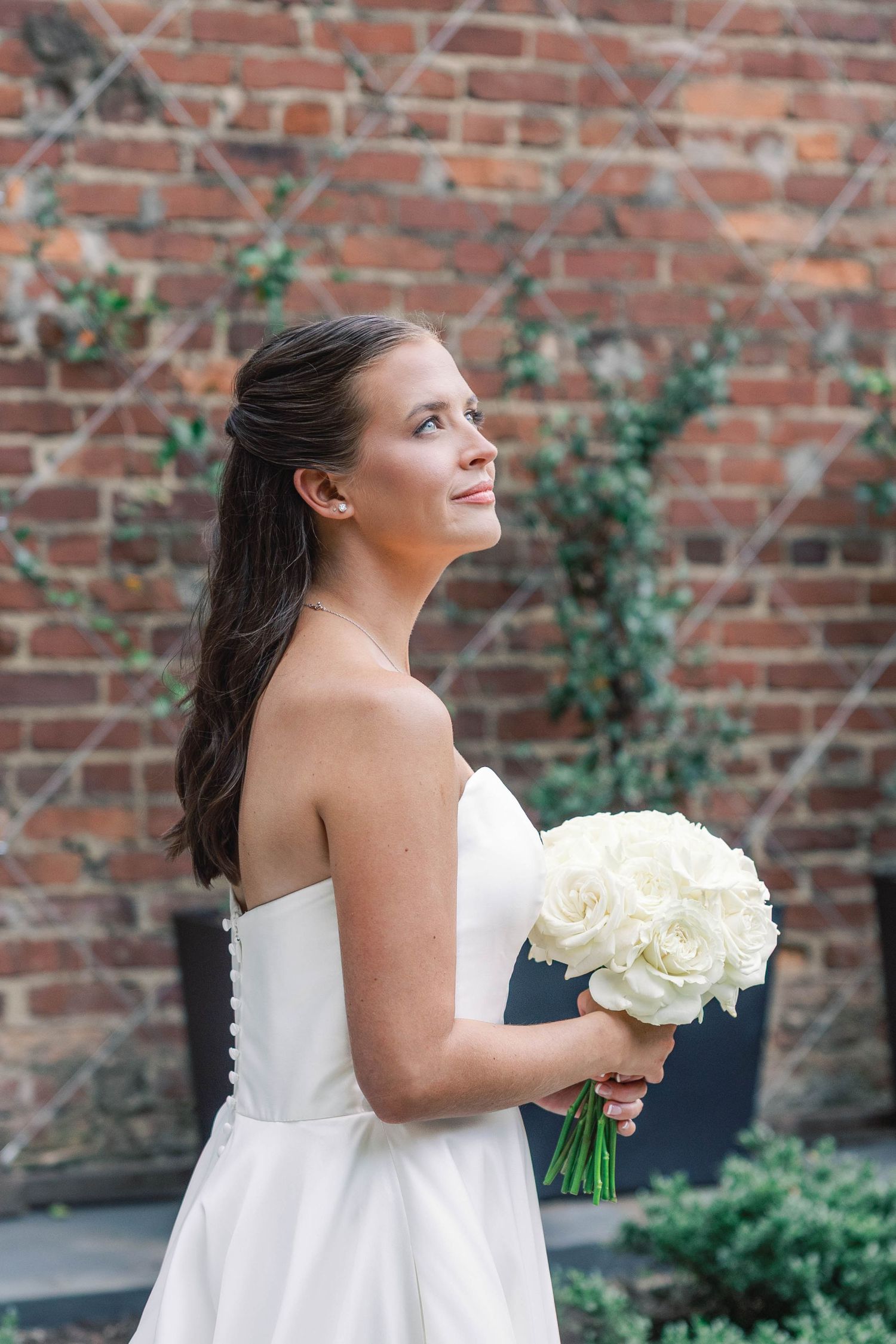 Bride in a white strapless wedding dress holds a bouquet of white roses against a rustic brick wall backdrop.