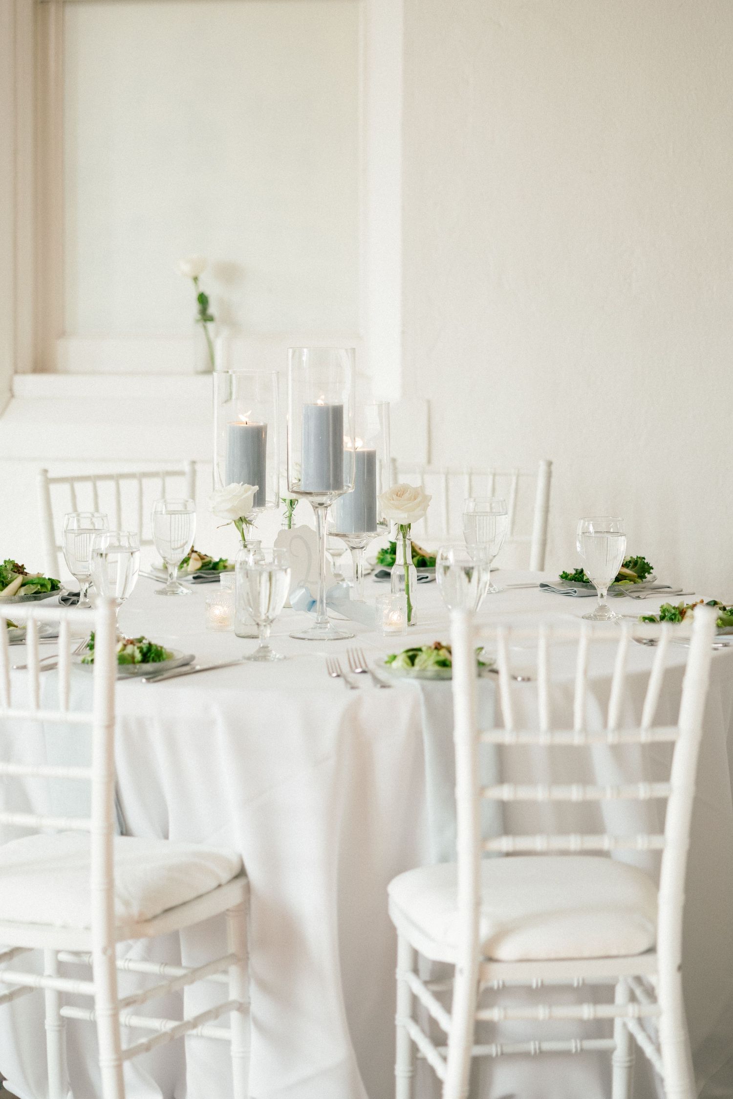 Elegant white wedding reception table setting with white chiavari chairs, tall gray candles and minimal floral accents.