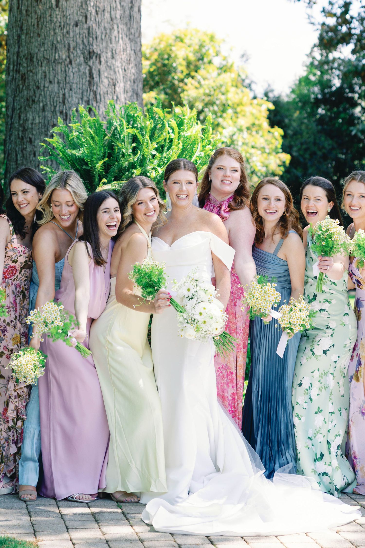 Wedding party of bridesmaids in pastel dresses gather around bride for outdoor photo.
