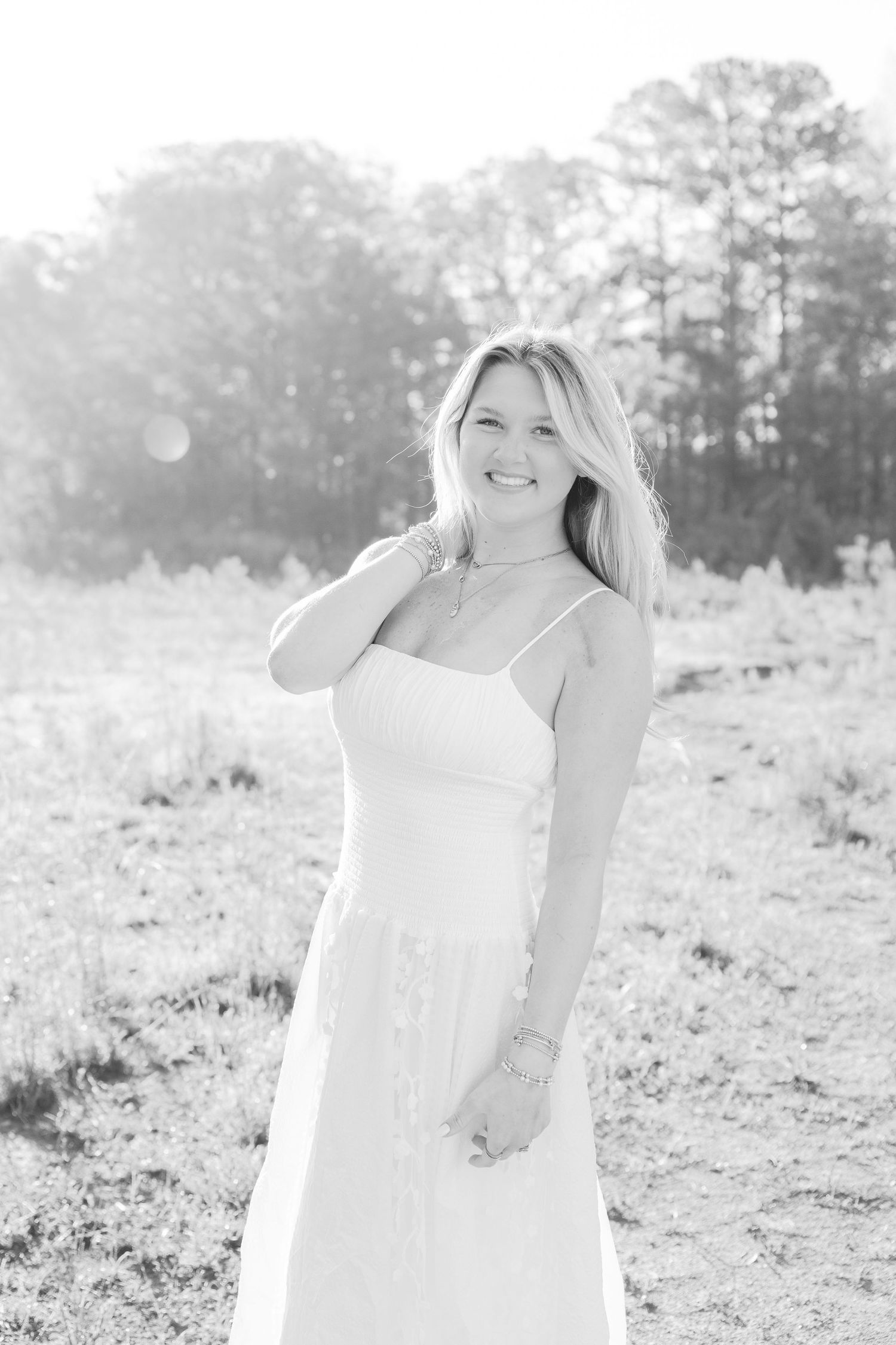 Woman in white dress smiles while posing in sunlit field with trees.