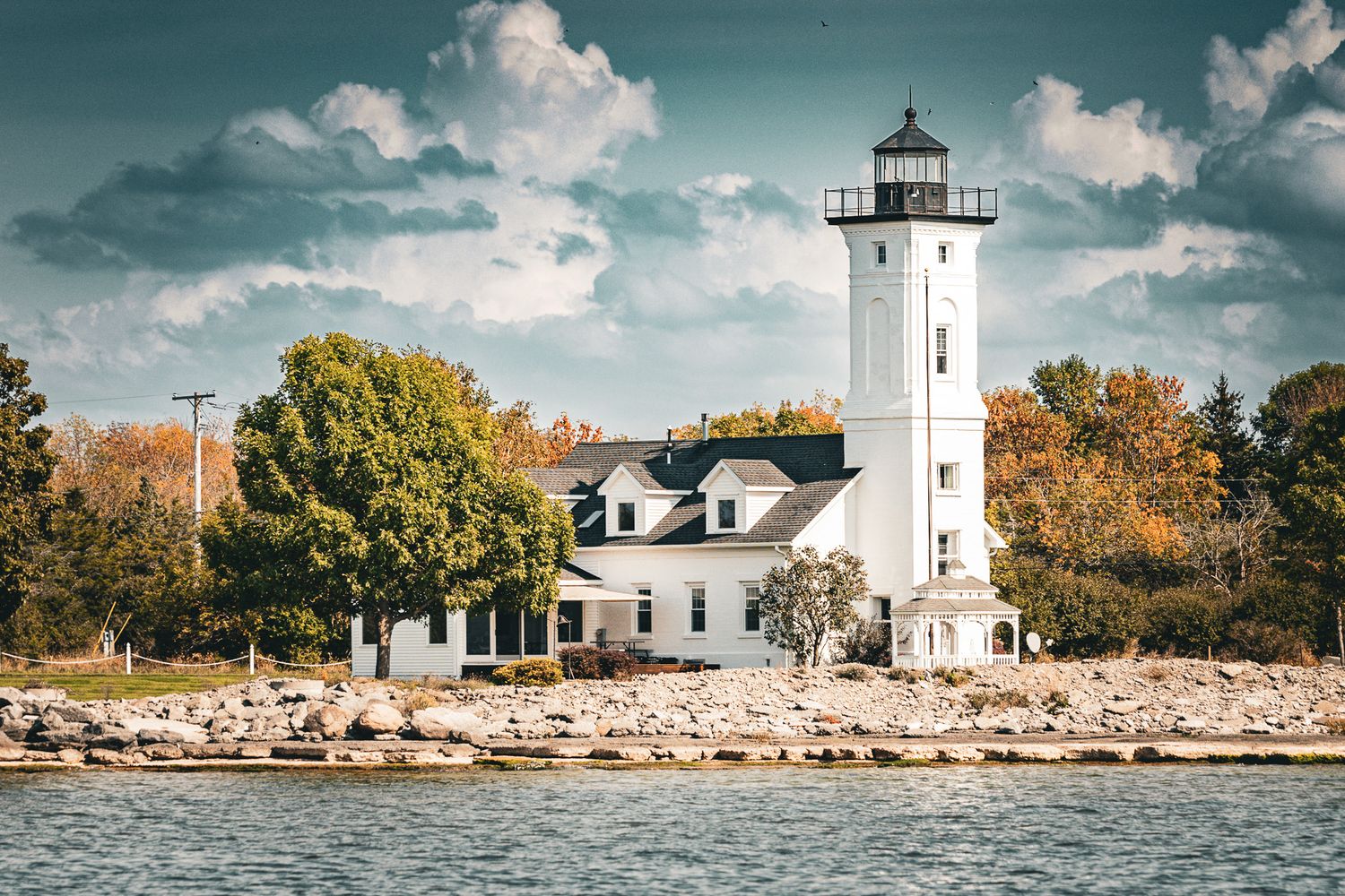 A white lighthouse and keeper's house stand on a rocky shoreline surrounded by autumn trees with teal waters below.